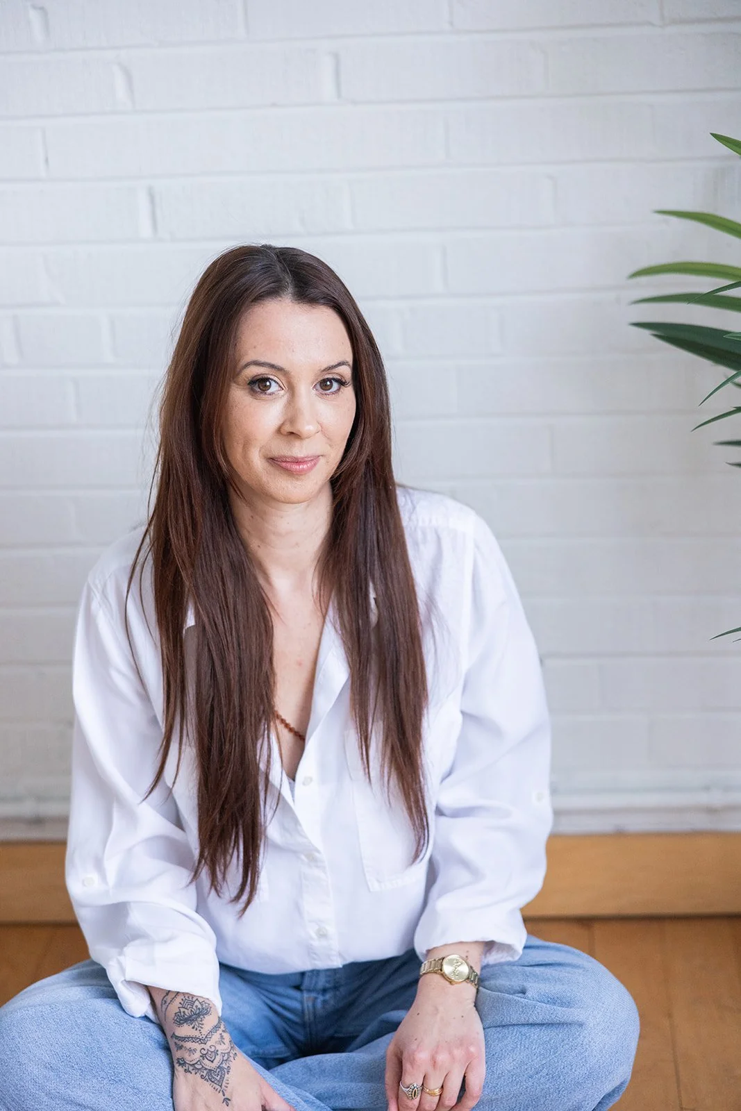A woman with long brown hair, wearing a white shirt and light blue pants, sitting on the floor with crossed legs, in front of a white brick wall with a green plant on her right.