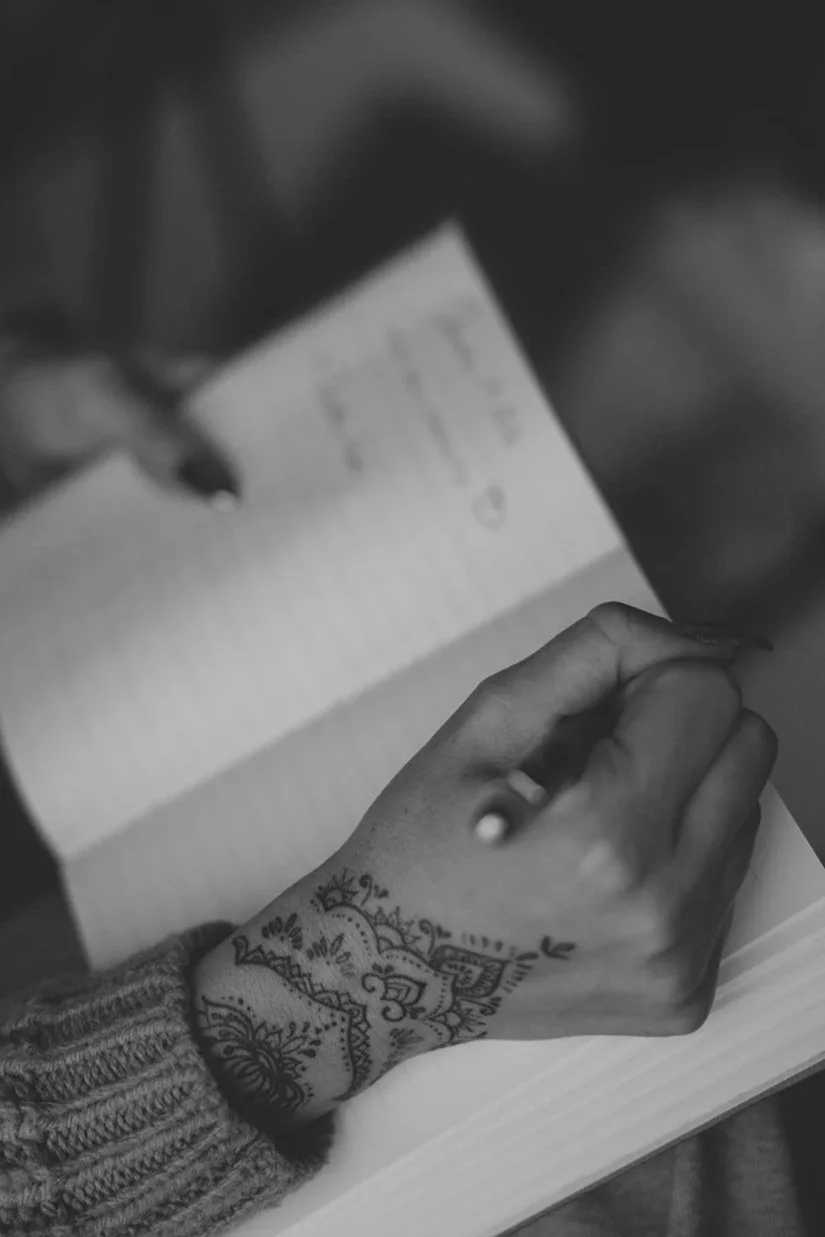 A person with a tattooed hand is writing or drawing on a piece of paper with a pen.