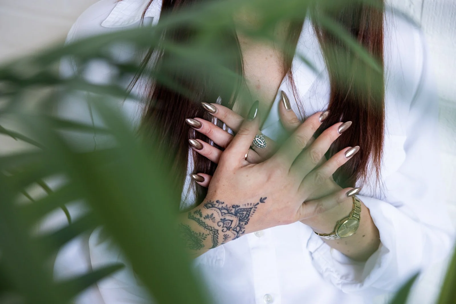 Close-up of a person's hands with metallic nails, jewelry, and tattoos, placed on their chest, partially obscured by green leaves in the foreground.