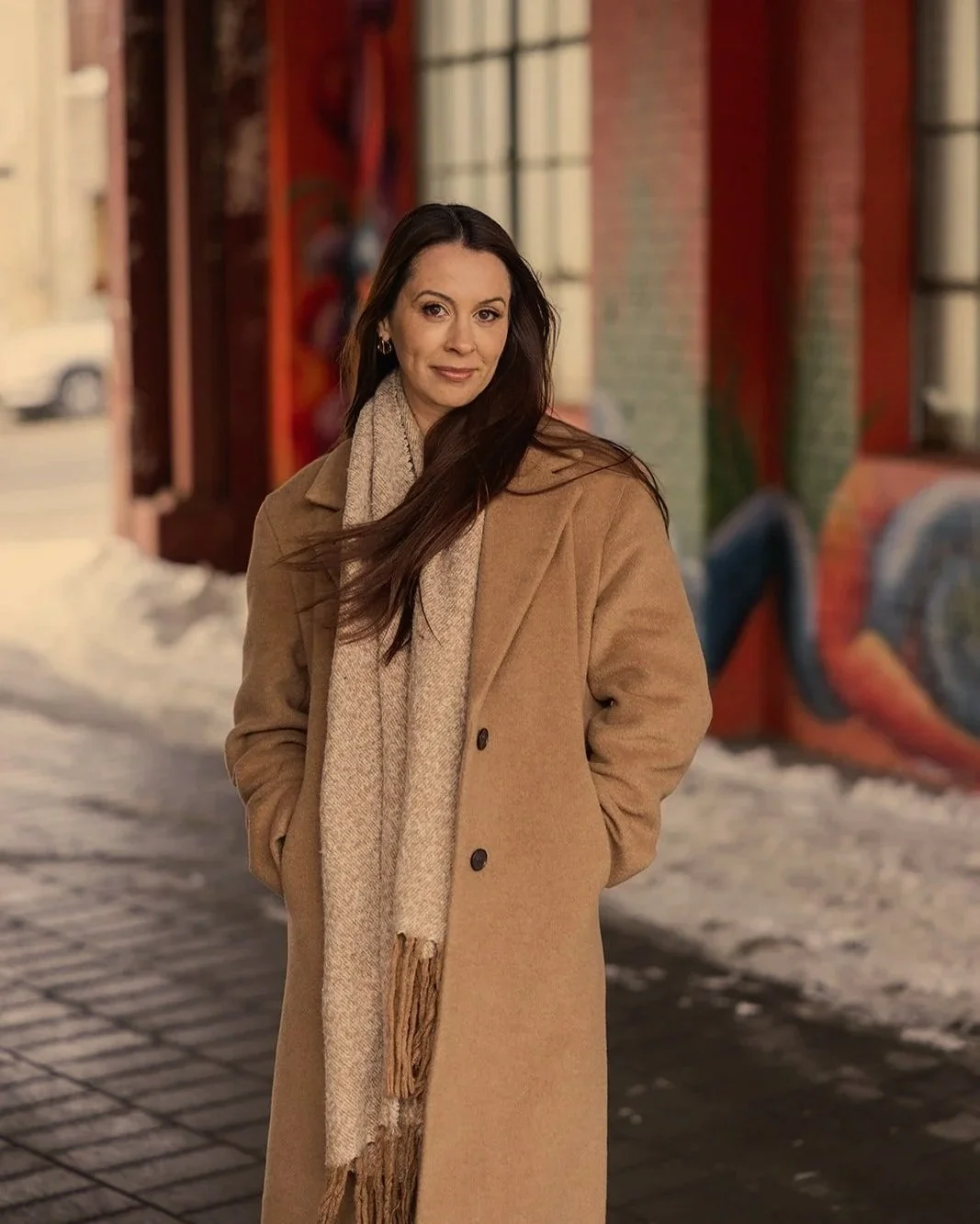A woman stands outdoors on a city street, wearing a tan coat and beige scarf, with colorful graffiti art in the background.