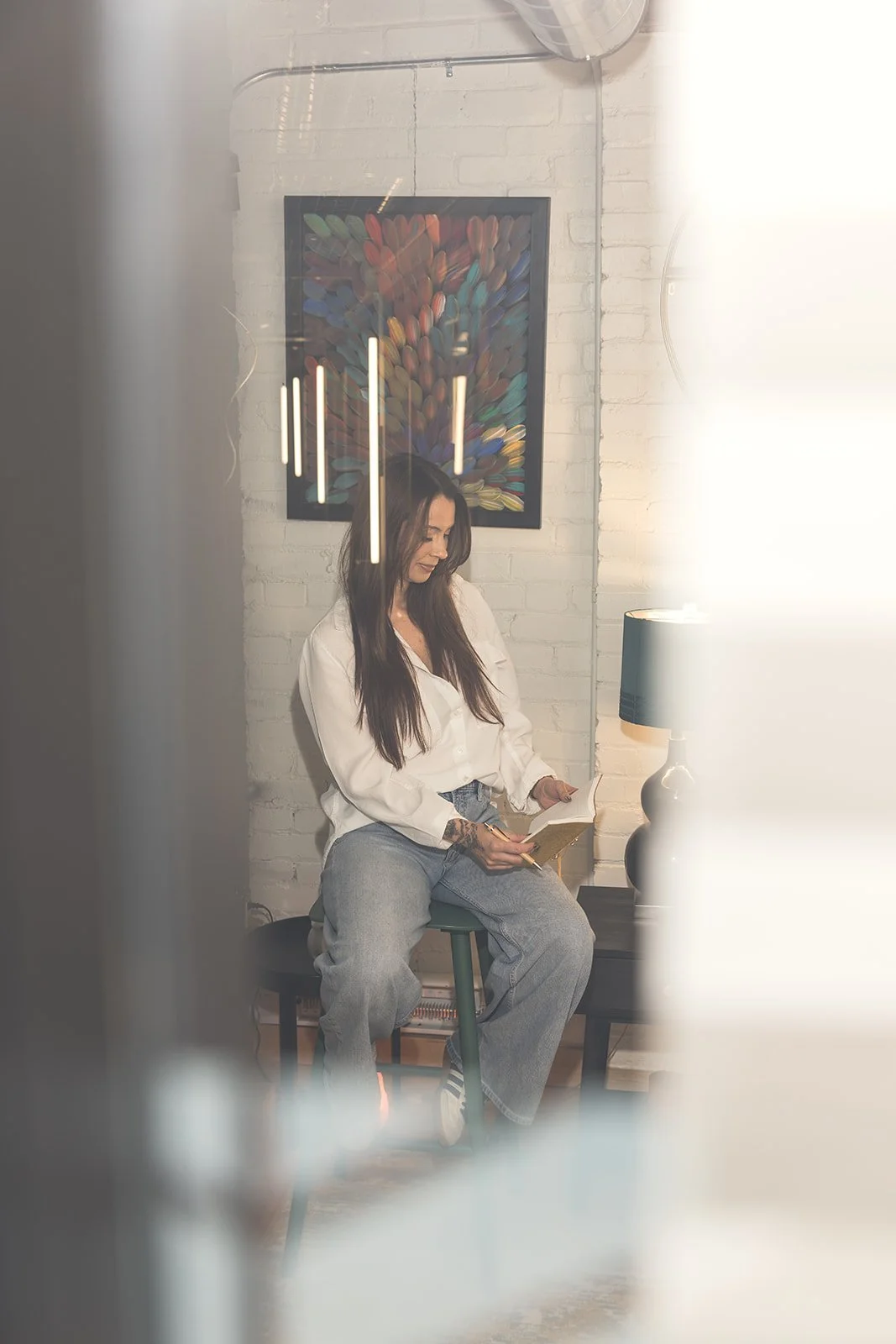 A woman with long dark hair sitting on a stool, reading a book in a room with a white brick wall, abstract artwork, and a lamp.