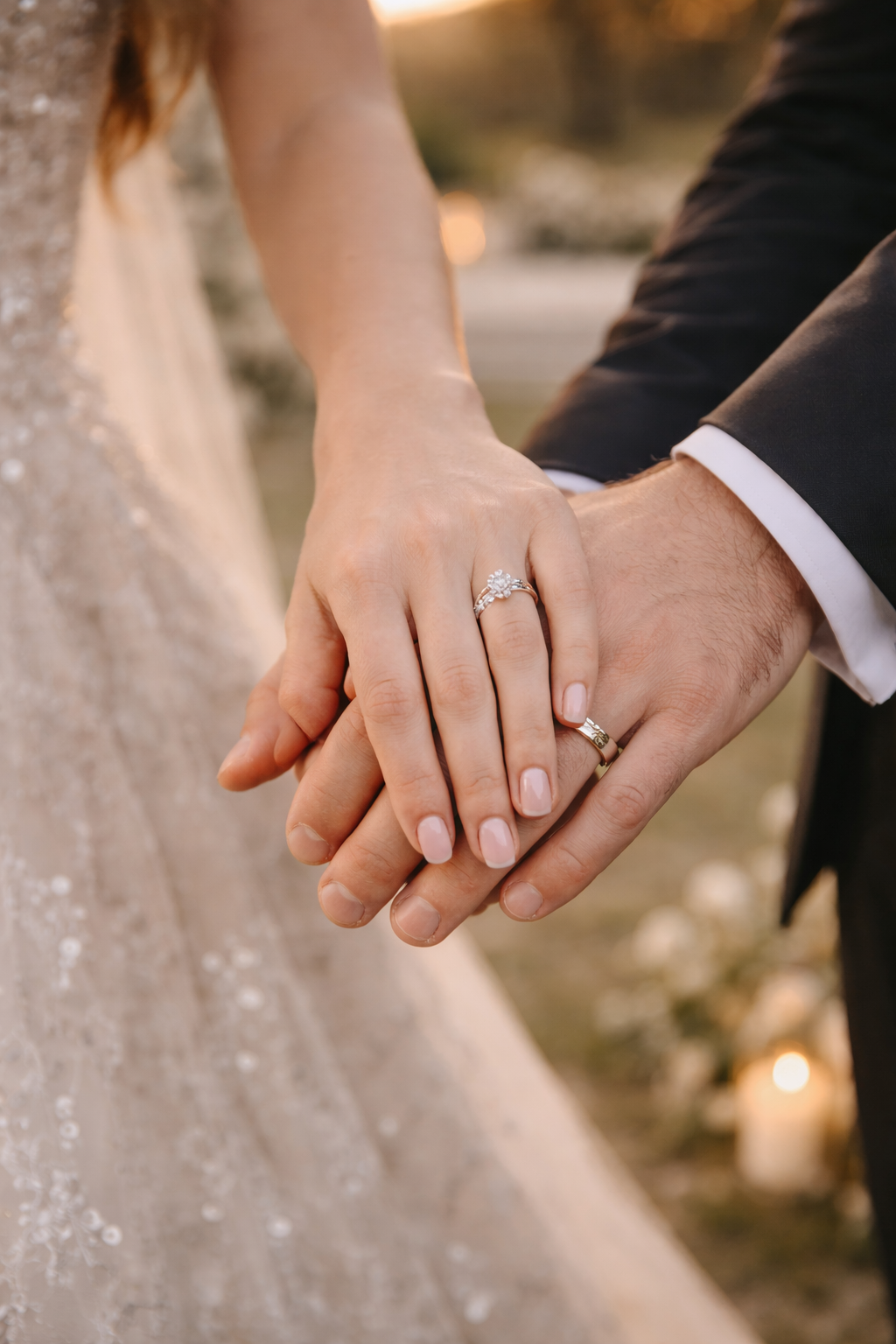 Close-up of a bride and groom holding hands, showing wedding rings. The bride's hand has a diamond engagement ring, and the groom's hand has a wedding band. The bride is wearing a cream-colored dress with sequin details, and the groom is wearing a black suit.