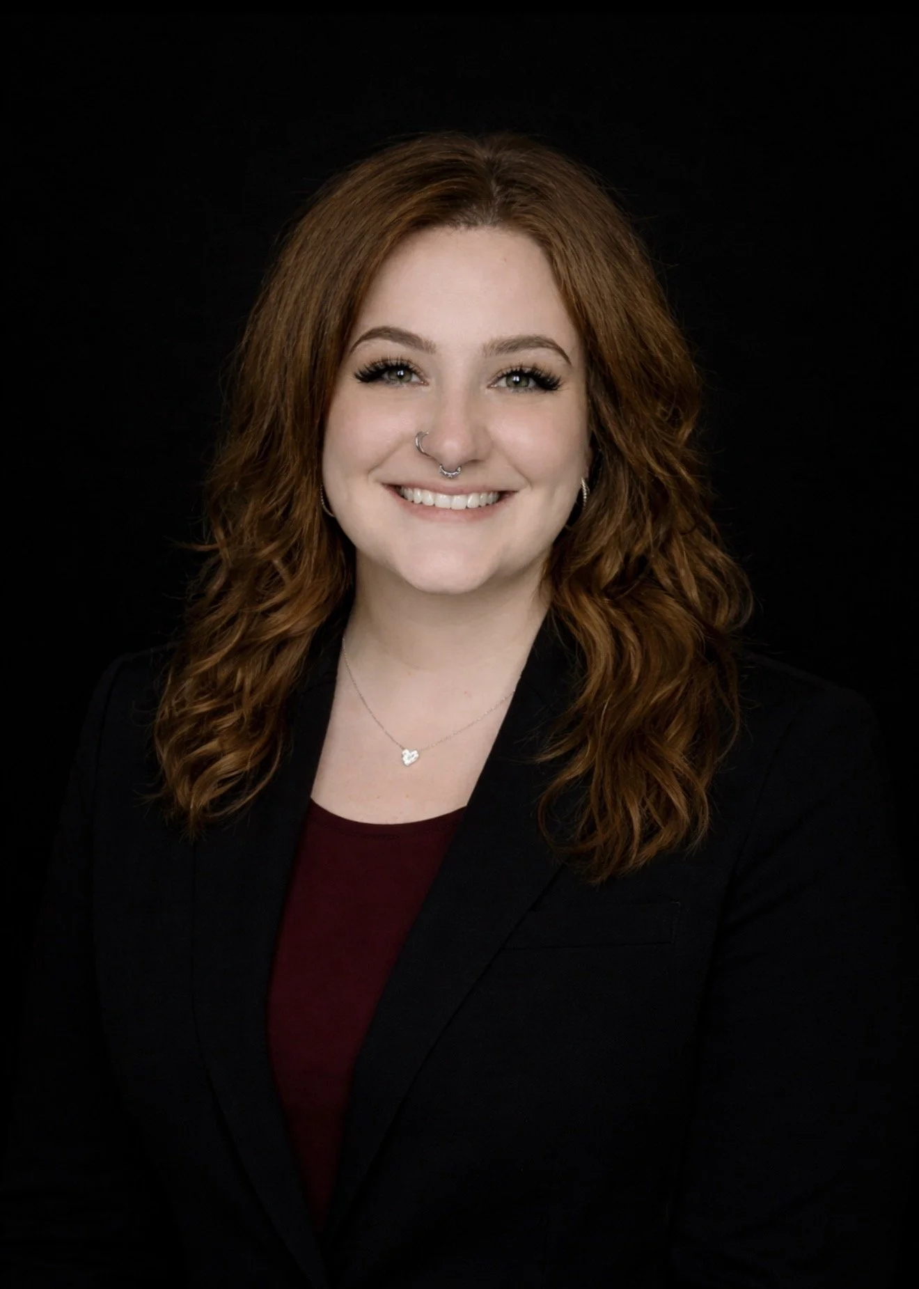 A woman with long, curly red hair smiling, wearing a black blazer, maroon top, and jewelry including a nose ring, earrings, and a necklace, against a black background.