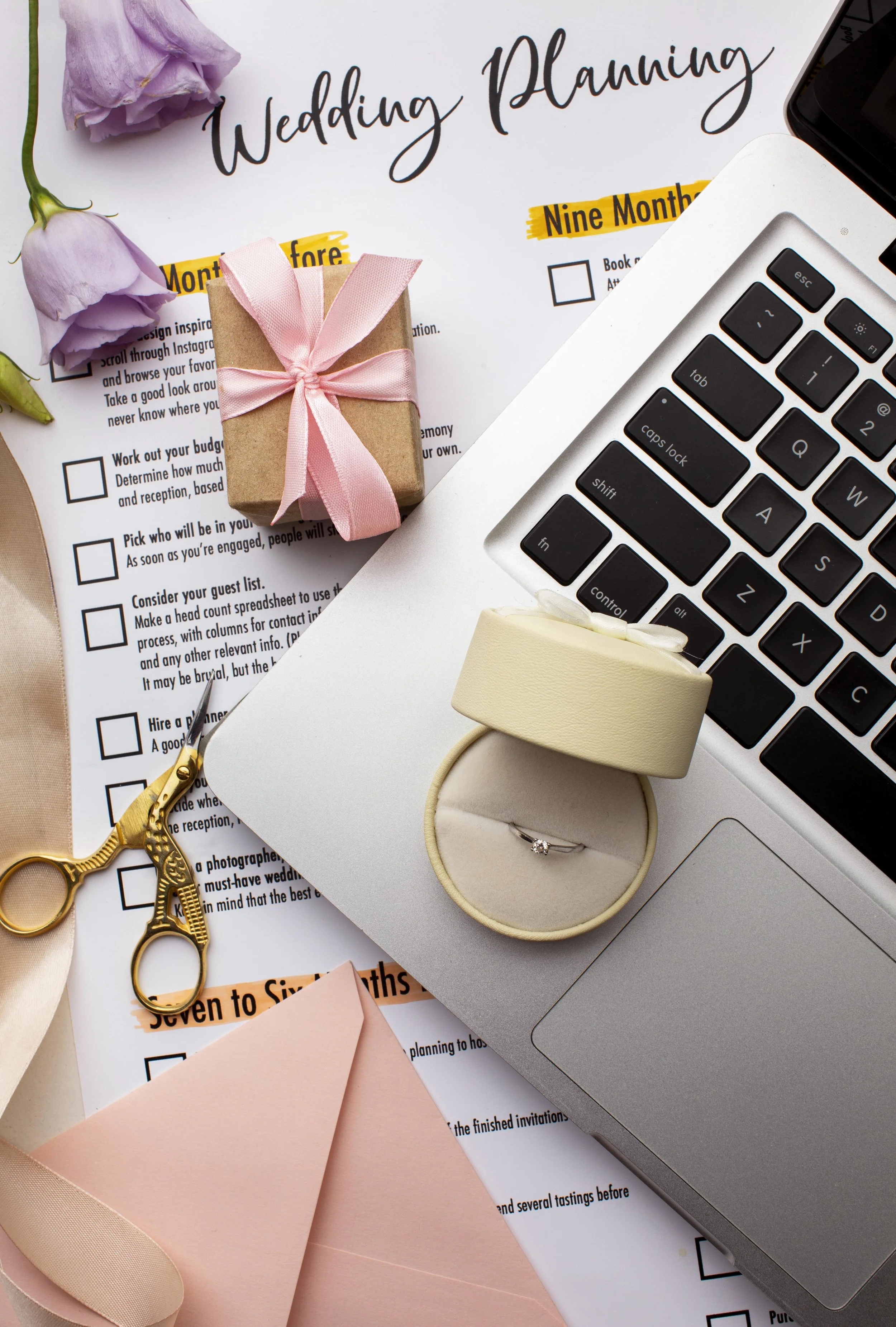 Wedding planning items on a desk, including a flower, a small gift box with pink ribbon, scissors, a ring in a beige ring box, a laptop, a wedding planning checklist, pink paper, and a smartphone.