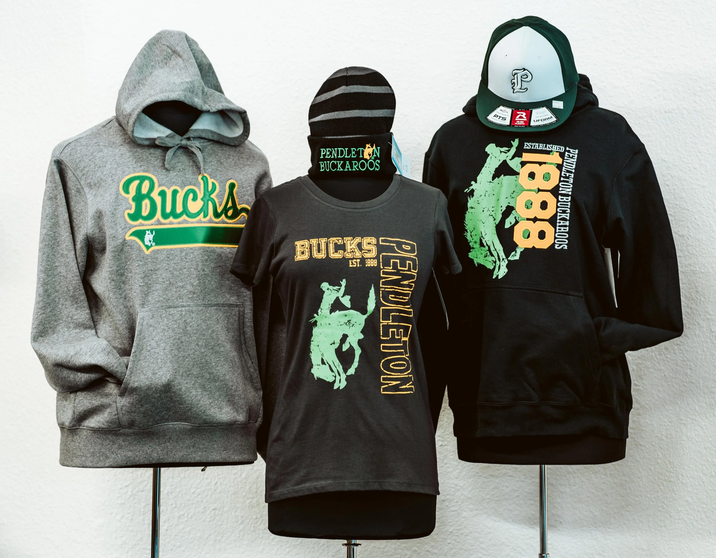 Three mannequin torsos displaying different Pendleton Bucks logo. The left wears a gray hooded sweatshirt, the middle wears a black T-shirt, and the right wears a black jacket and a black and white baseball cap with a logo.