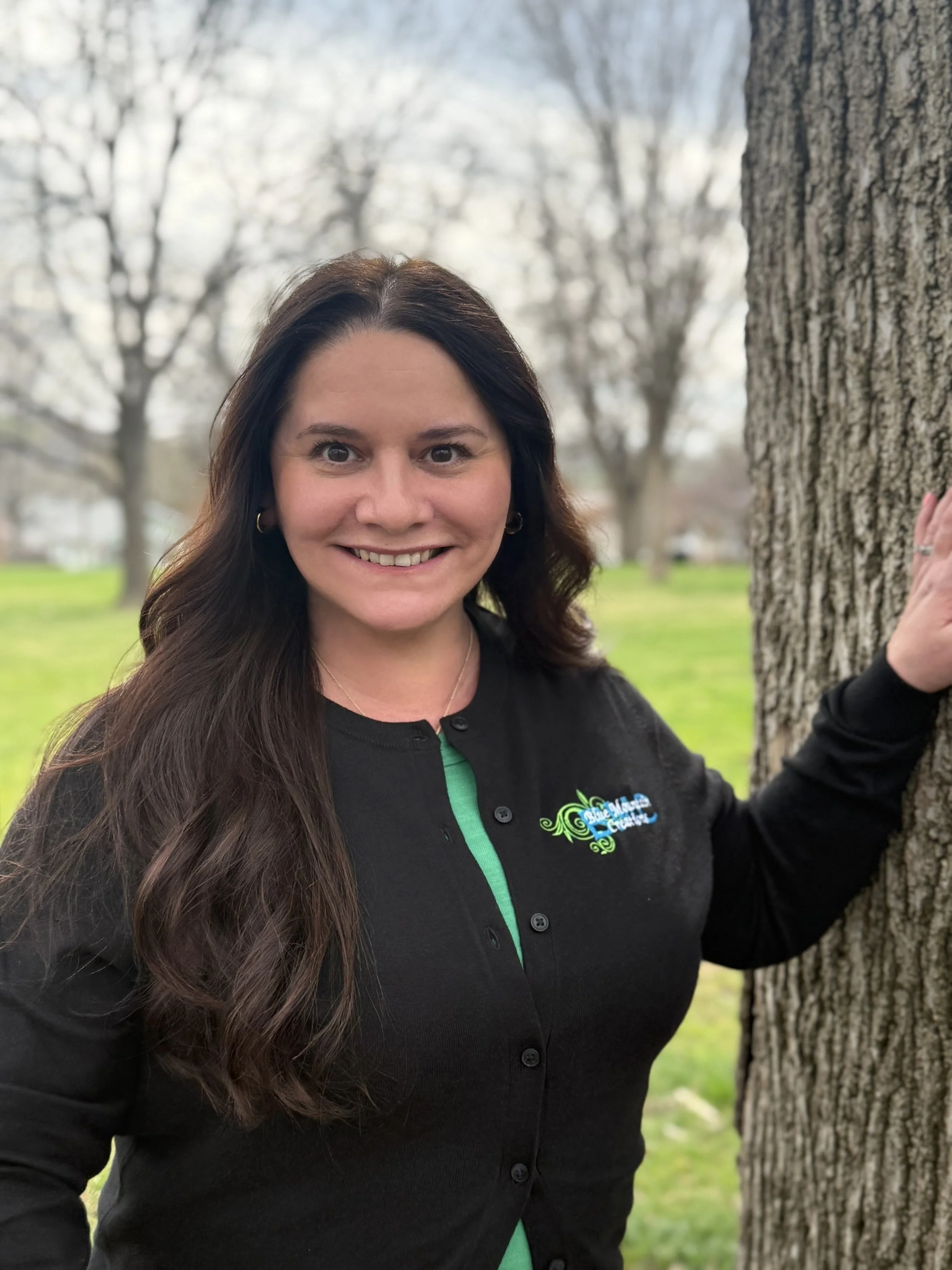 Woman with long brown hair smiling, standing outdoors in front of trees, wearing a black jacket with a Blue Mountain Creations logo.