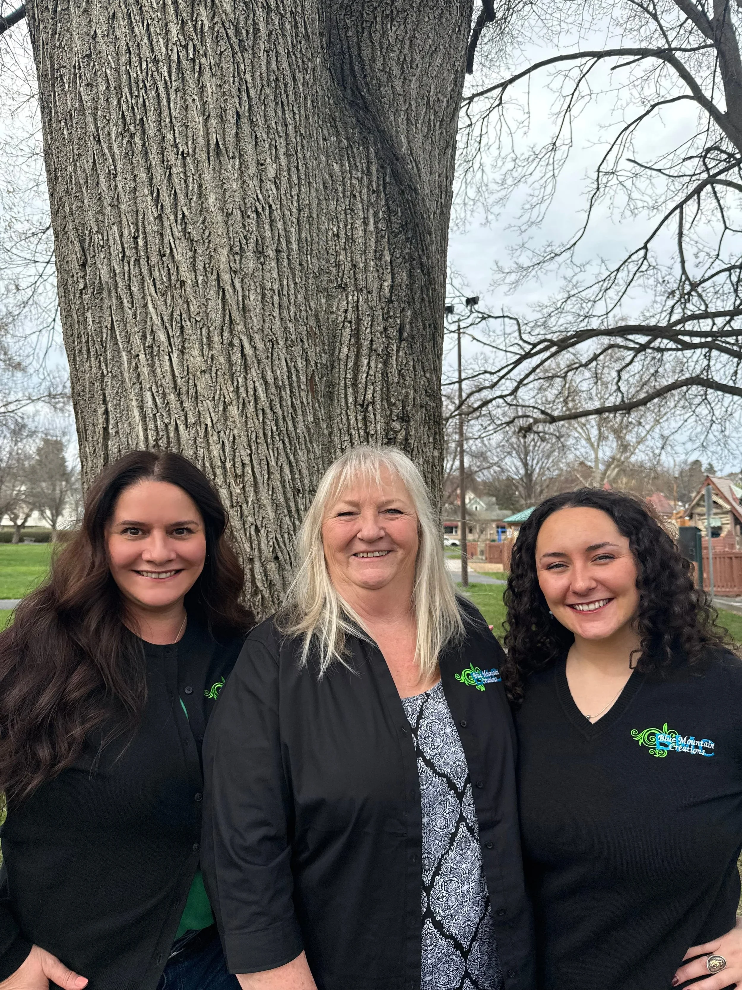 Three women standing outdoors in front of a large tree, smiling at the camera, with a park and houses in the background.