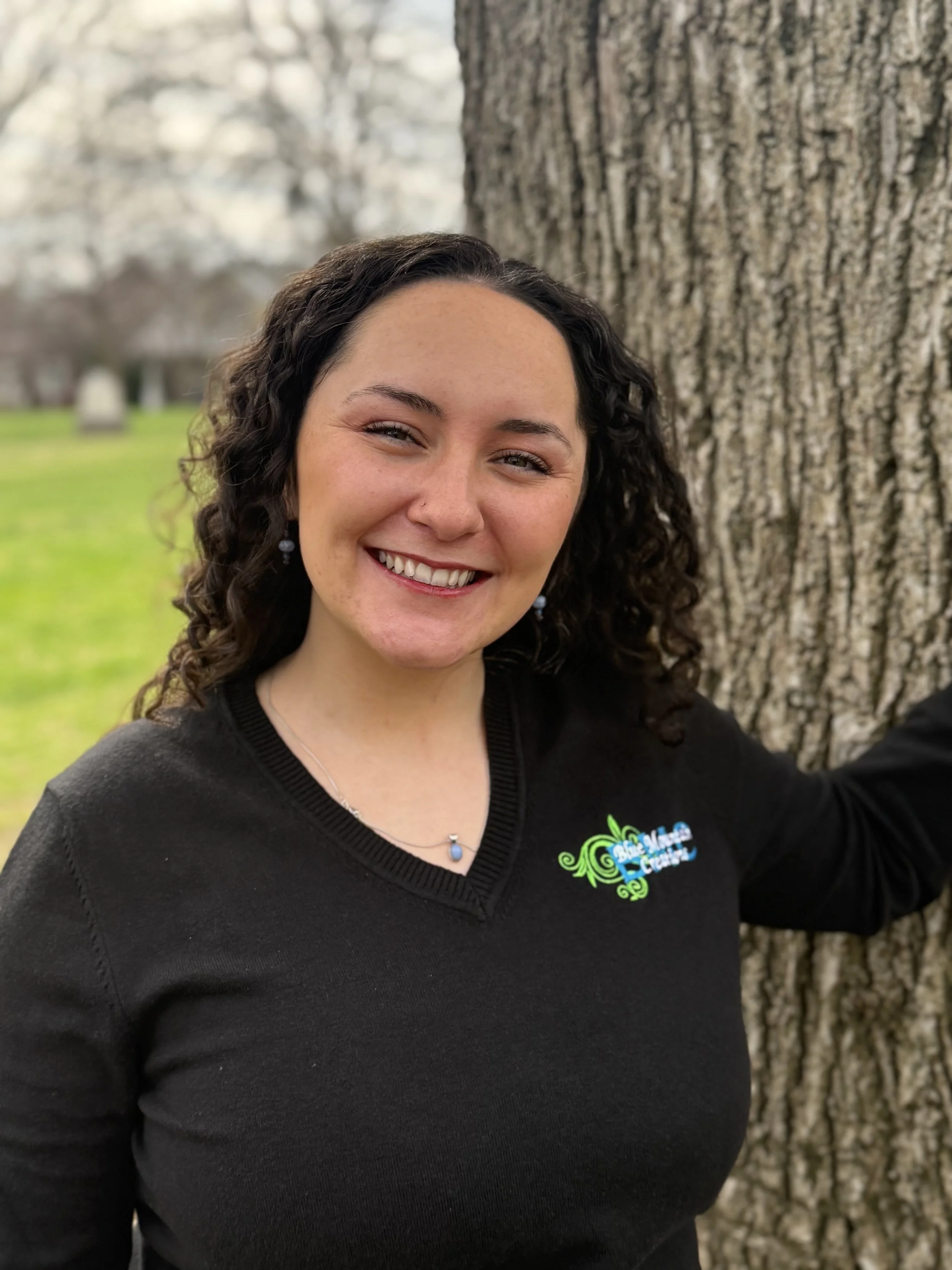 A woman with curly dark hair smiling, standing outdoors next to a large tree trunk, wearing a black top with a Blue Mountain Creations logo.