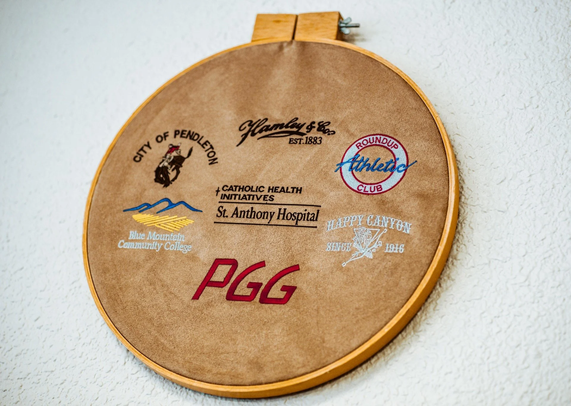 Wooden plaque mounted on wall with various logos and text, including 'City of Pendleton,' 'Hamley & Co. Est. 1883,' 'Catholic Health Initiatives,' 'St. Anthony Hospital,' and others.
