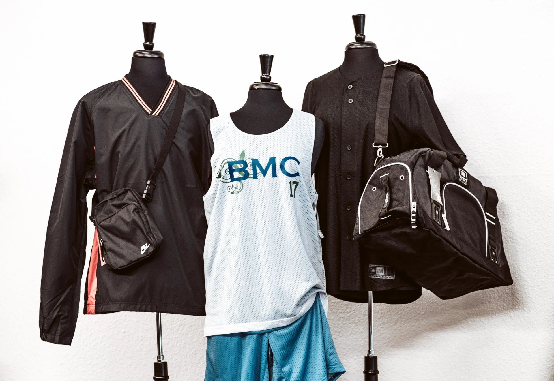 Display of sportswear on mannequins, including a black jacket with Nike bag, a white basketball jersey with 'BMC' logo, and a black button-up shirt with backpack.
