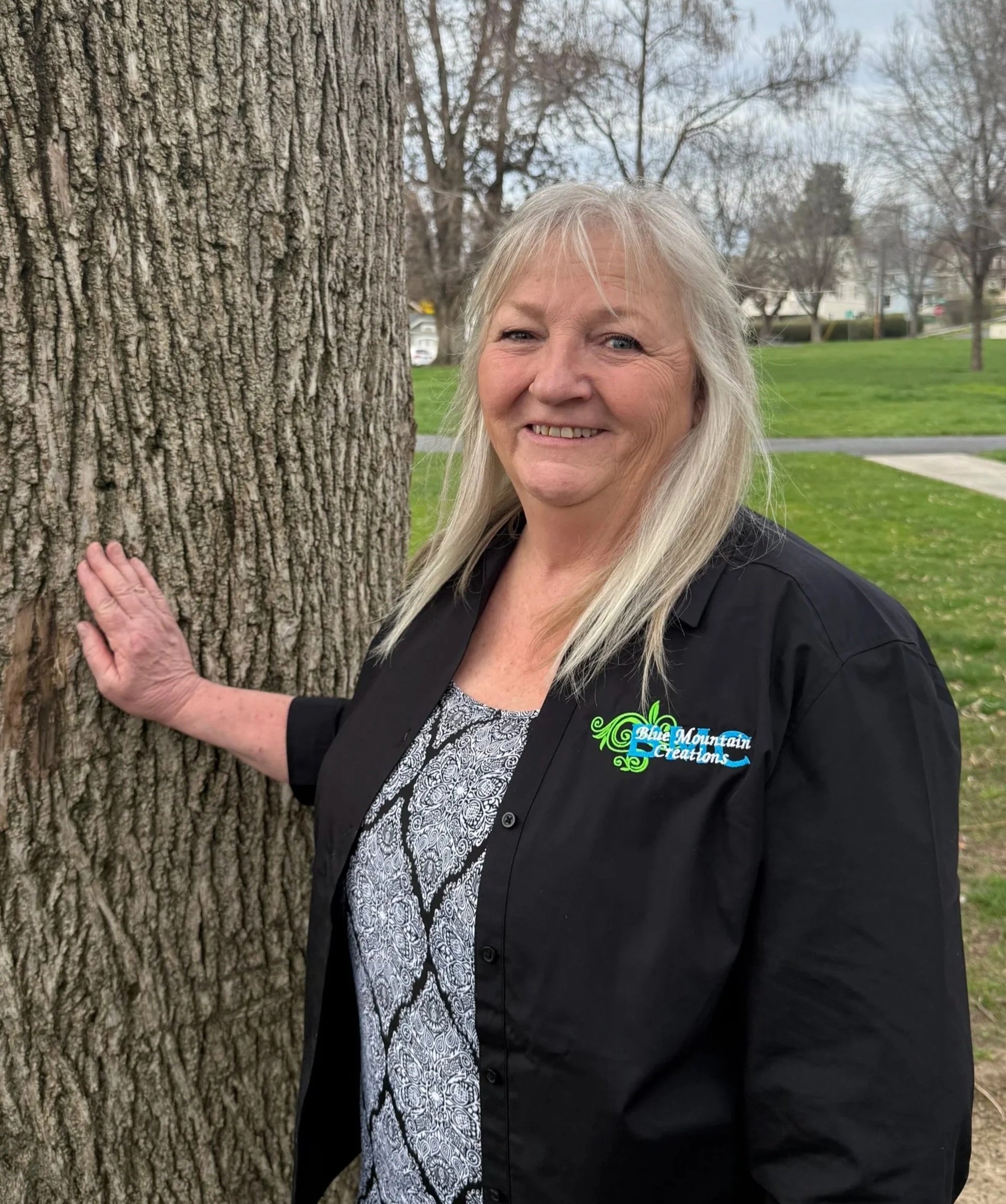 A smiling woman with long gray hair, wearing a black jacket with a logo that says 'Blue Mountain Creations.' She is standing outdoors next to a large tree, with a park and trees in the background on a cloudy day.