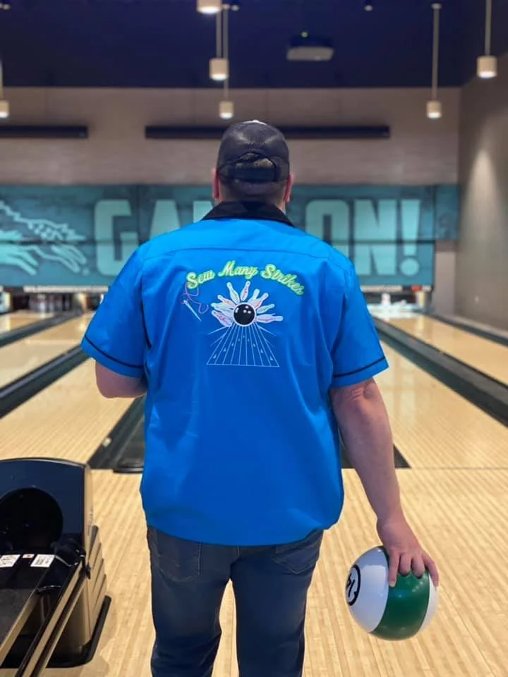 Person in a blue bowling shirt and backwards cap holding a bowling ball in a bowling alley.