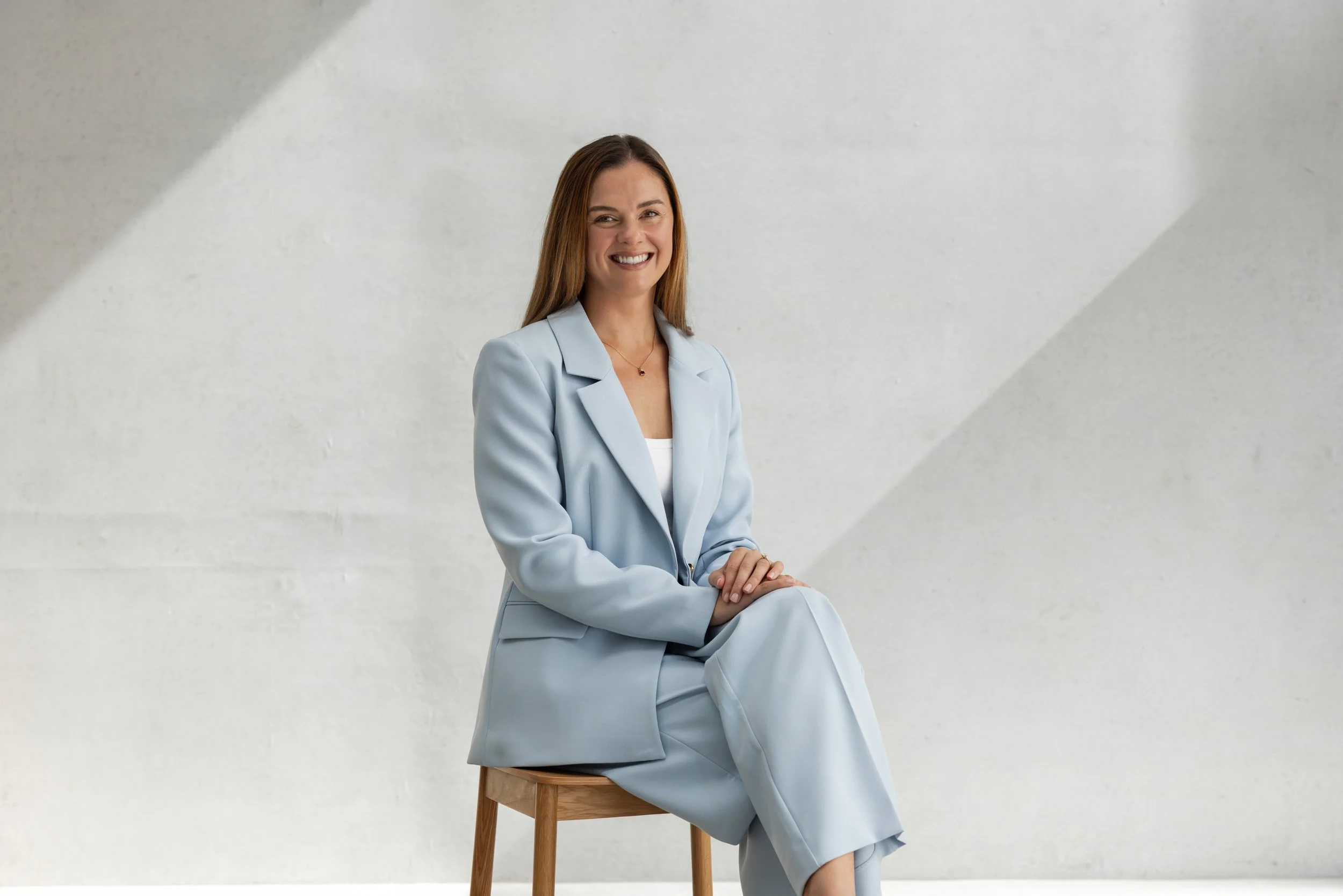 A smiling woman with long brown hair in a light blue suit sitting on a wooden stool against a white wall background.