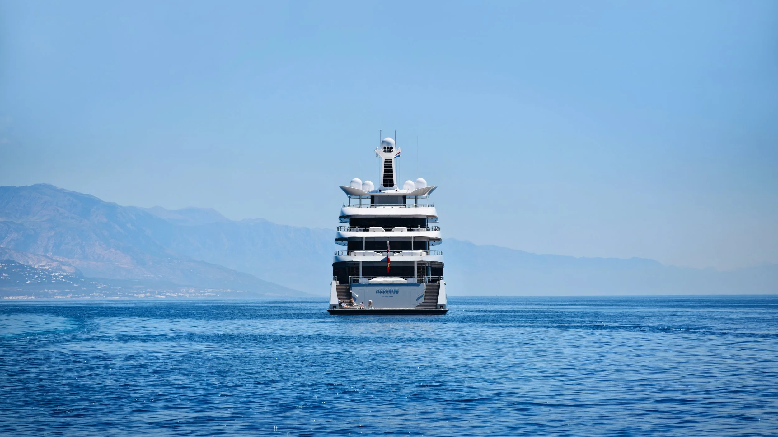 A large white luxury cruise ship sailing on calm blue water with mountains in the background under a clear blue sky.