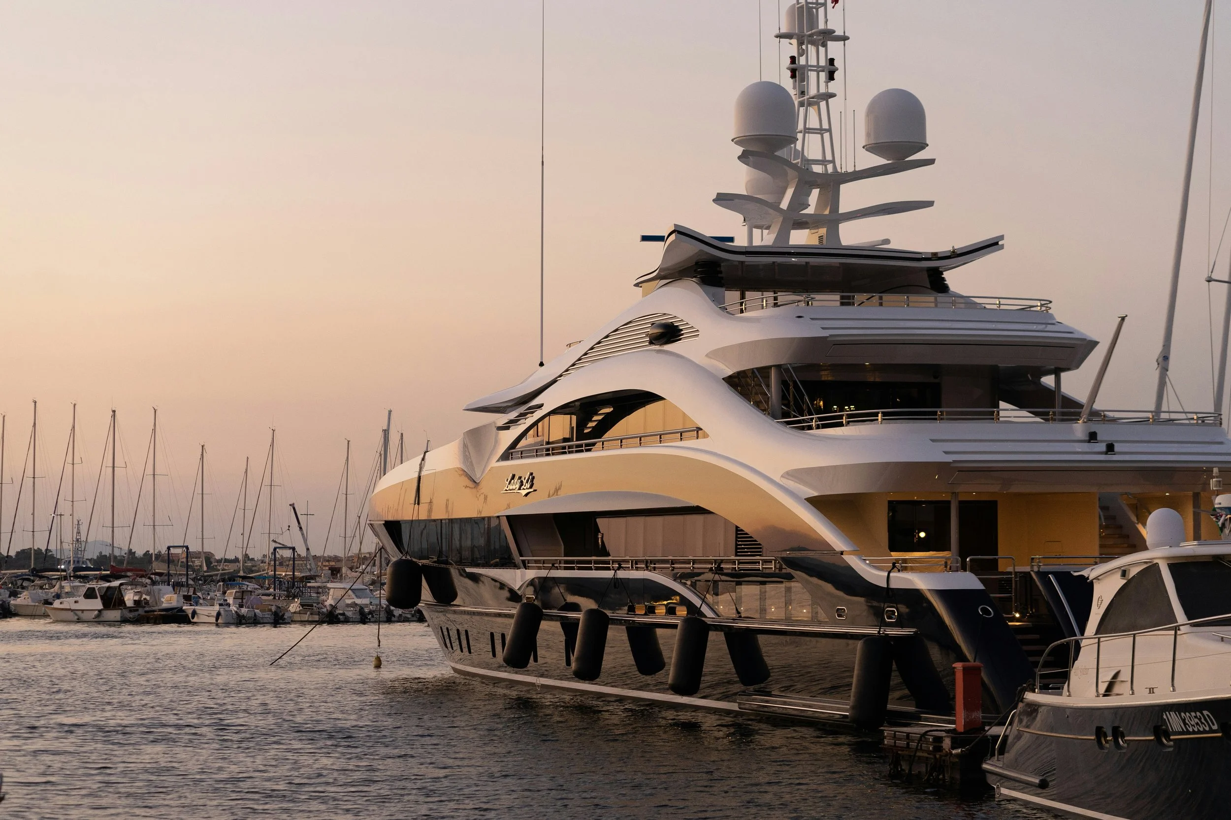 A large luxury yacht docked in a marina during sunset, with smaller boats and sailboats visible in the background.