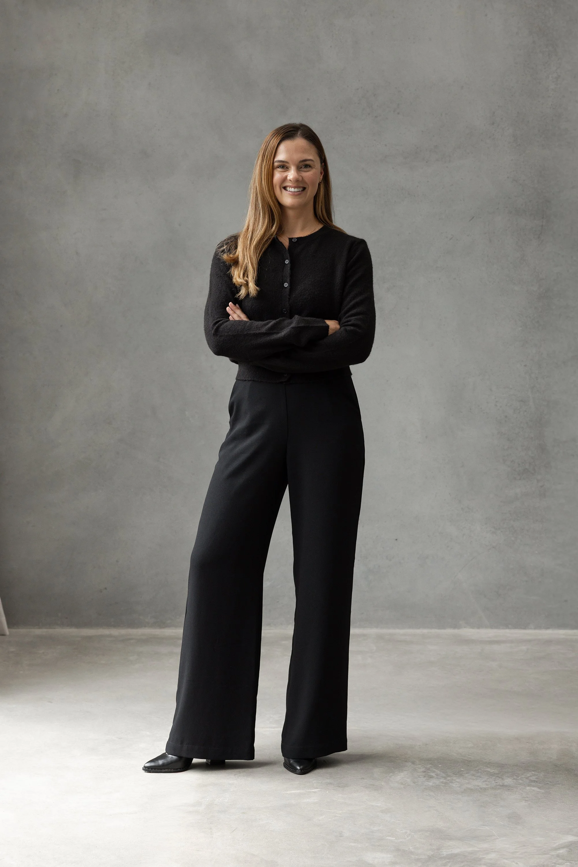 Woman with long brown hair wearing black sweater and wide-leg black pants standing in front of a neutral gray background, smiling with arms crossed.