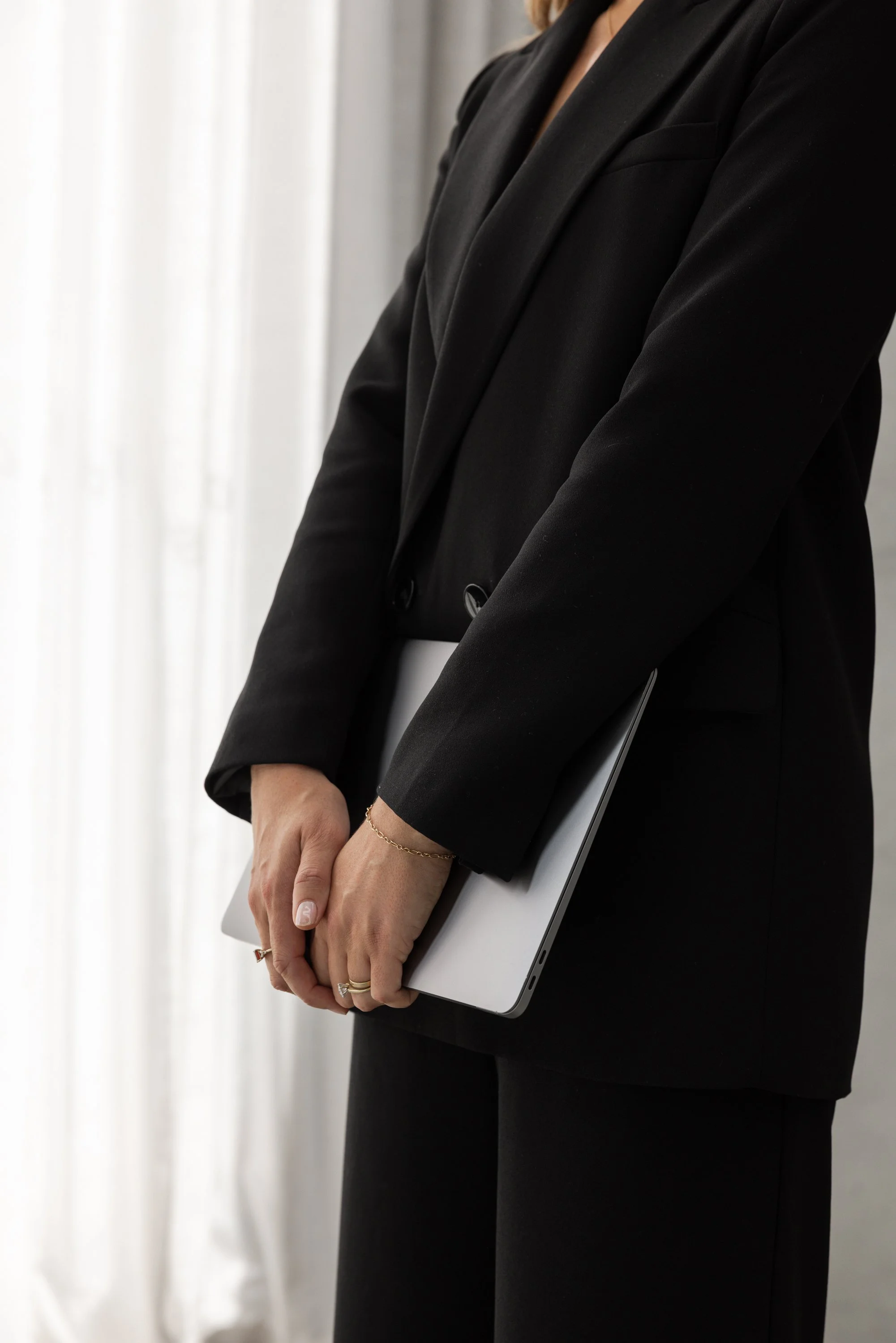 A shorebased superyacht purser dressed in black holding a closed laptop with both hands, standing near white curtains.