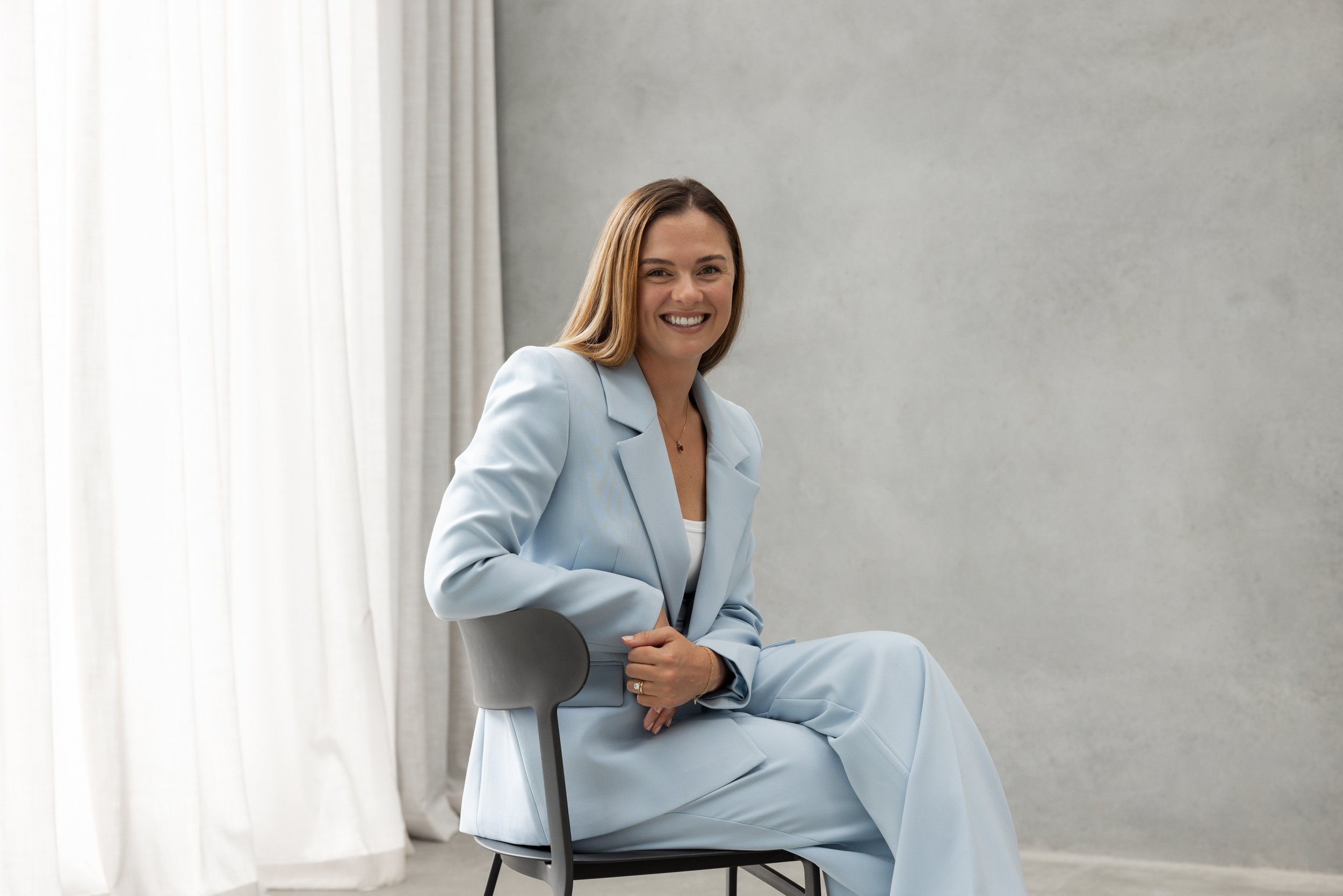 A shoreside superyacht purser in a light blue suit sitting on a chair, smiling, in front of a gray wall with white curtains.
