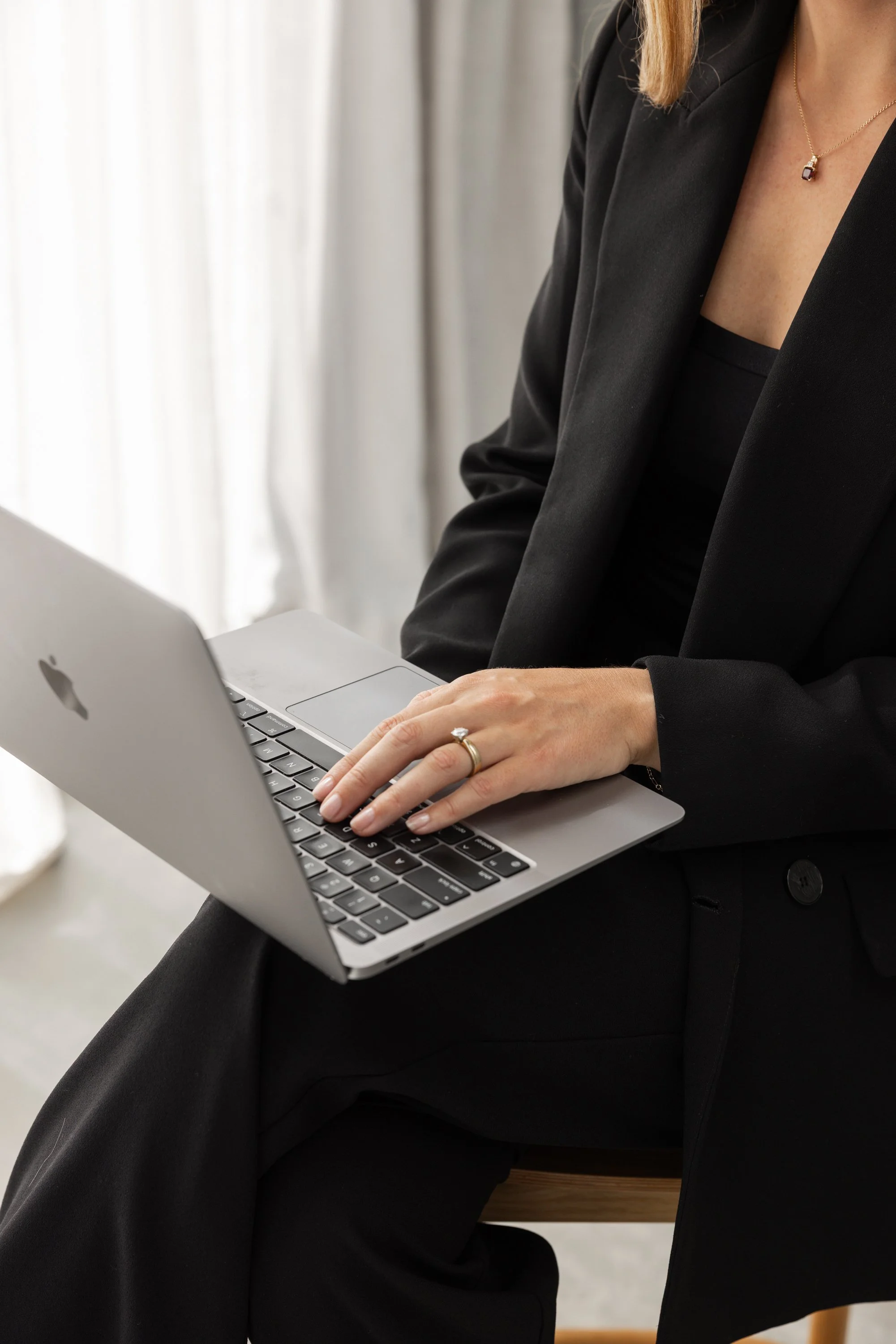 A woman sitting on a wooden chair wearing a black blazer and black clothing, using a silver MacBook laptop placed on her lap.