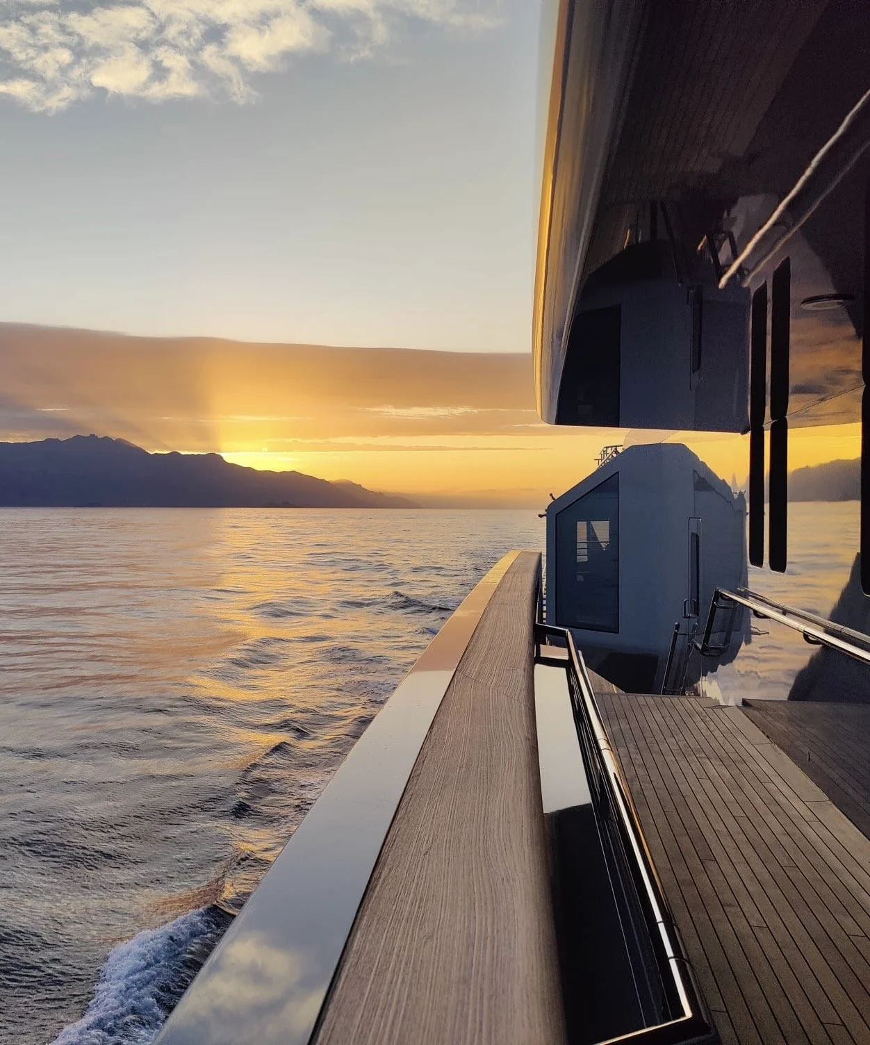 A photo of a yacht at sea during sunset, showing the side of the yacht, the water, and a mountainous horizon in the distance.