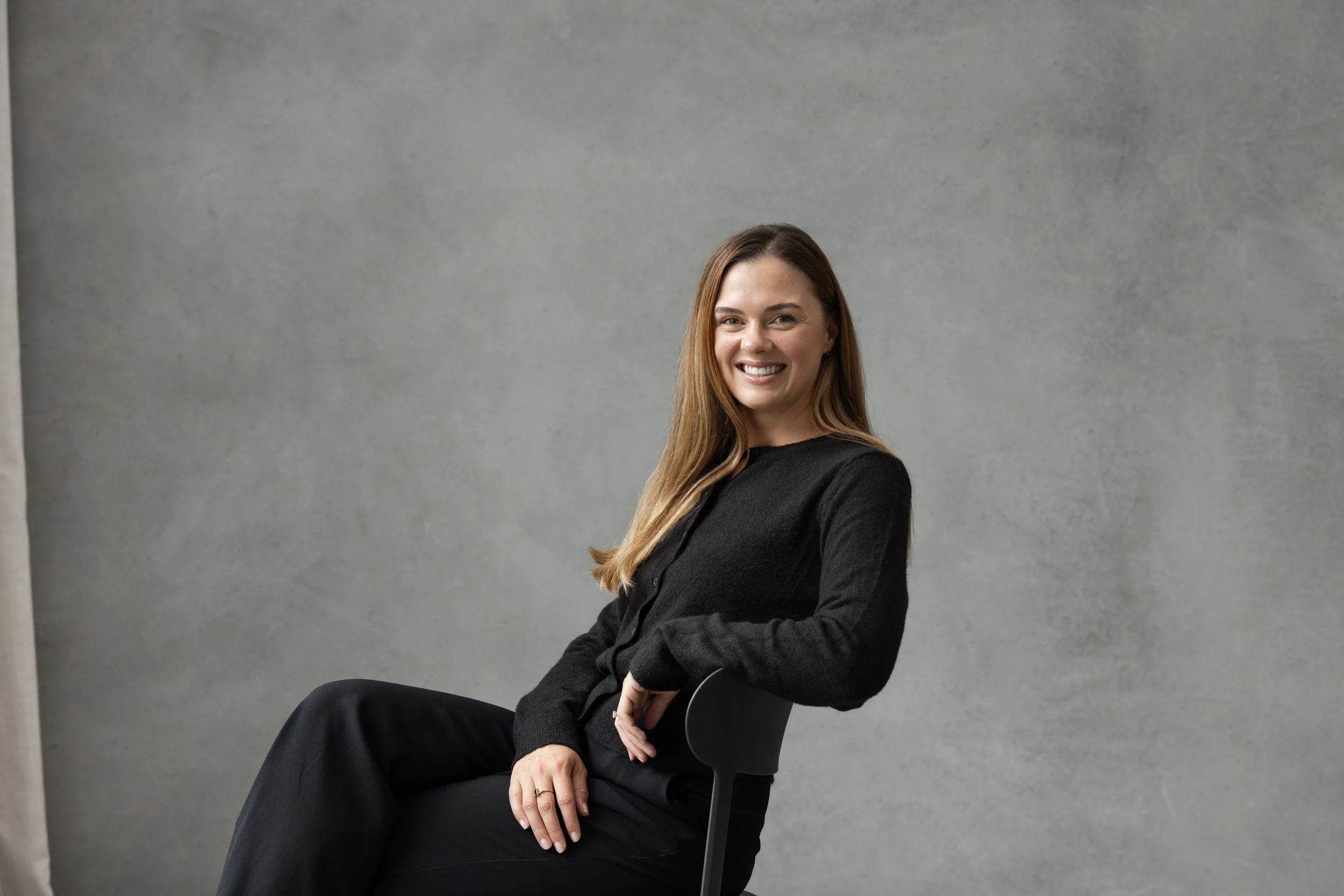 A remote superyacht purser with long brown hair, smiling, sitting on a black chair against a plain grey wall, wearing a black long-sleeve top and black pants.