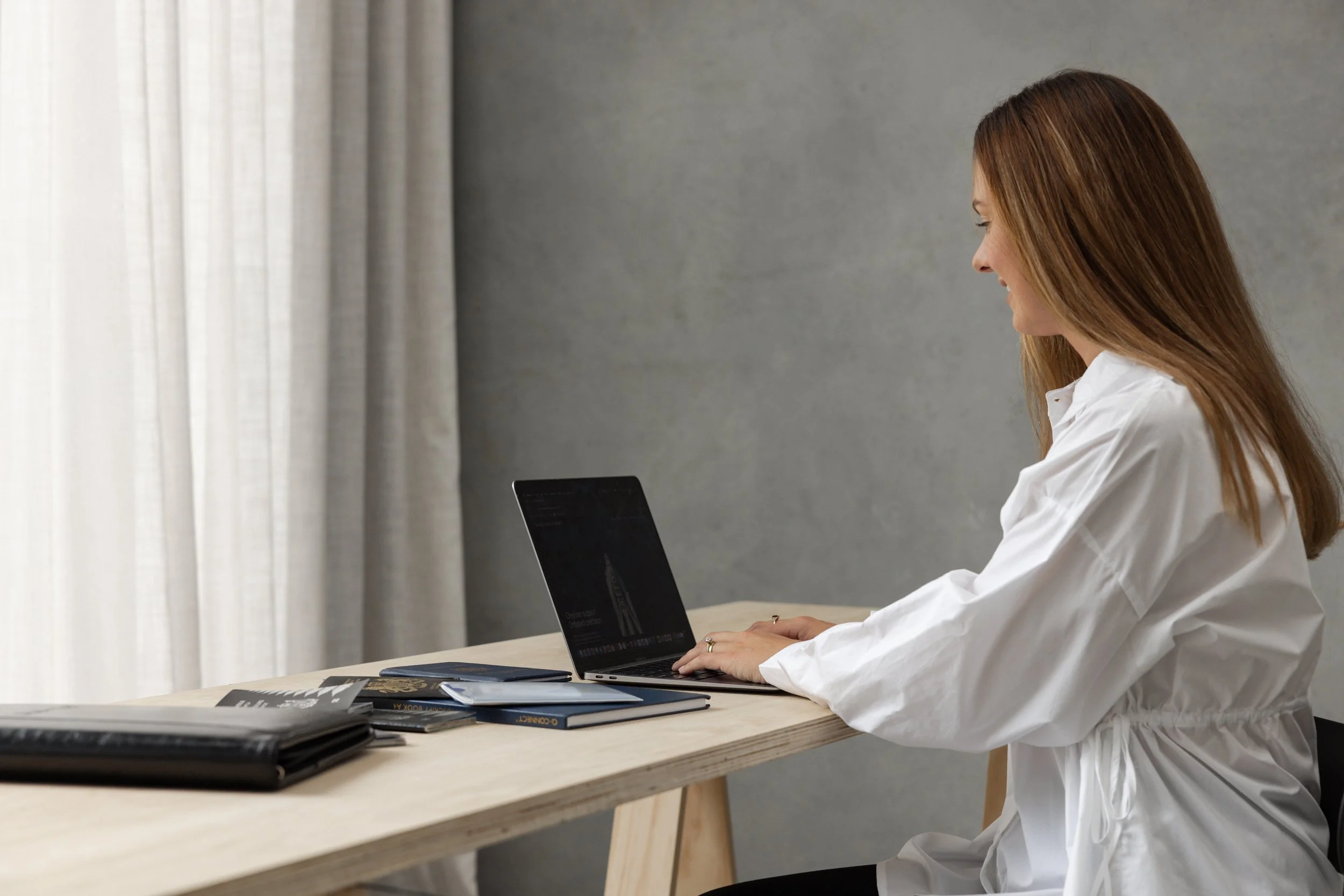 A woman with long brown hair sitting at a light wooden table working on a laptop, with a gray wall and curtains in the background.