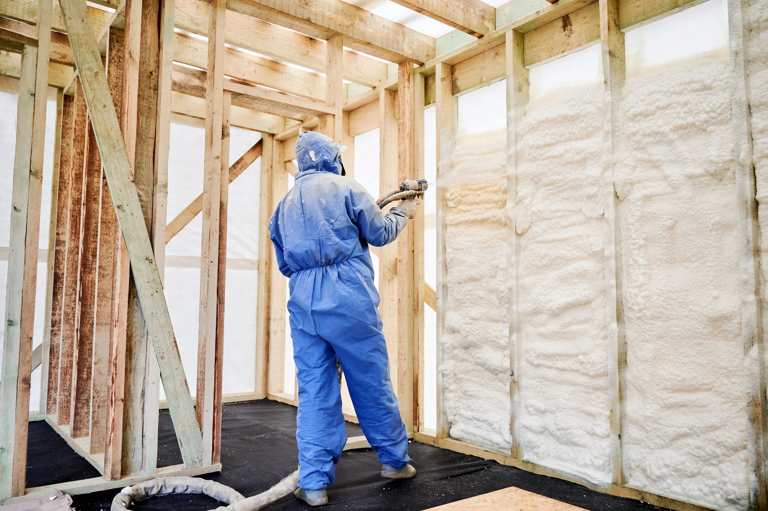 Worker in blue protective suit spray foaming wall insulation inside a wooden framed building under construction.