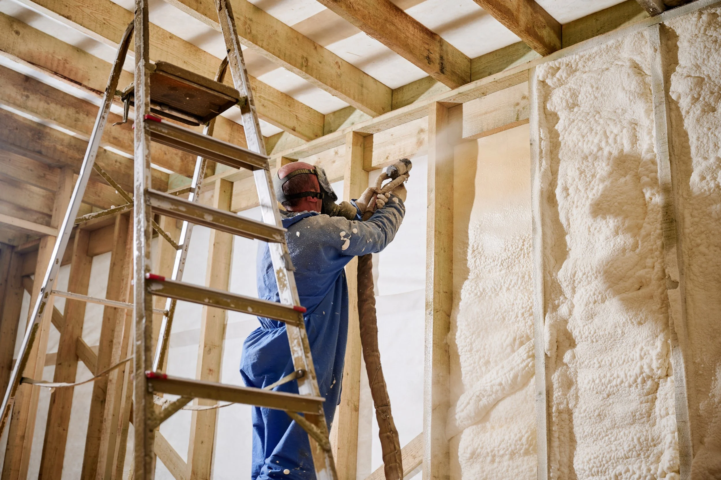 A construction worker wearing a mask and gloves installs insulation spray foam between wooden studs in a building under construction. A ladder is positioned nearby.