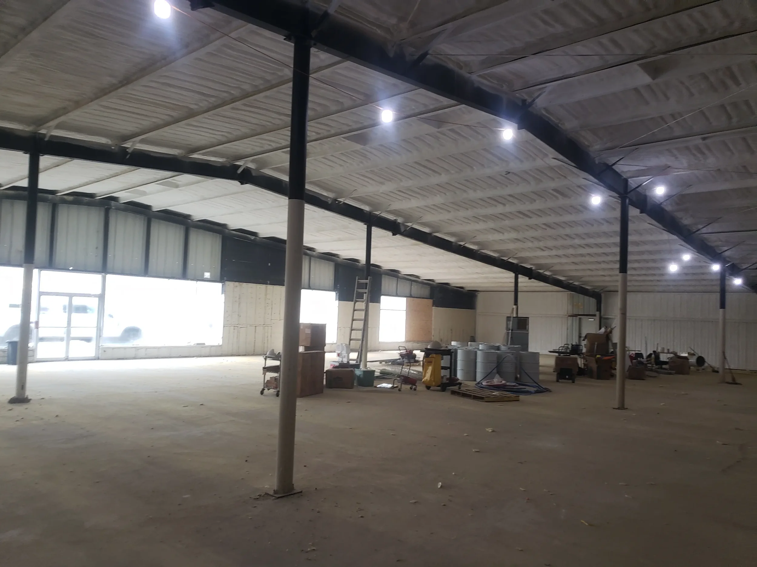 Empty indoor space under construction with steel supports, a ladder, and construction tools, with a curved ceiling and large windows at the far end.