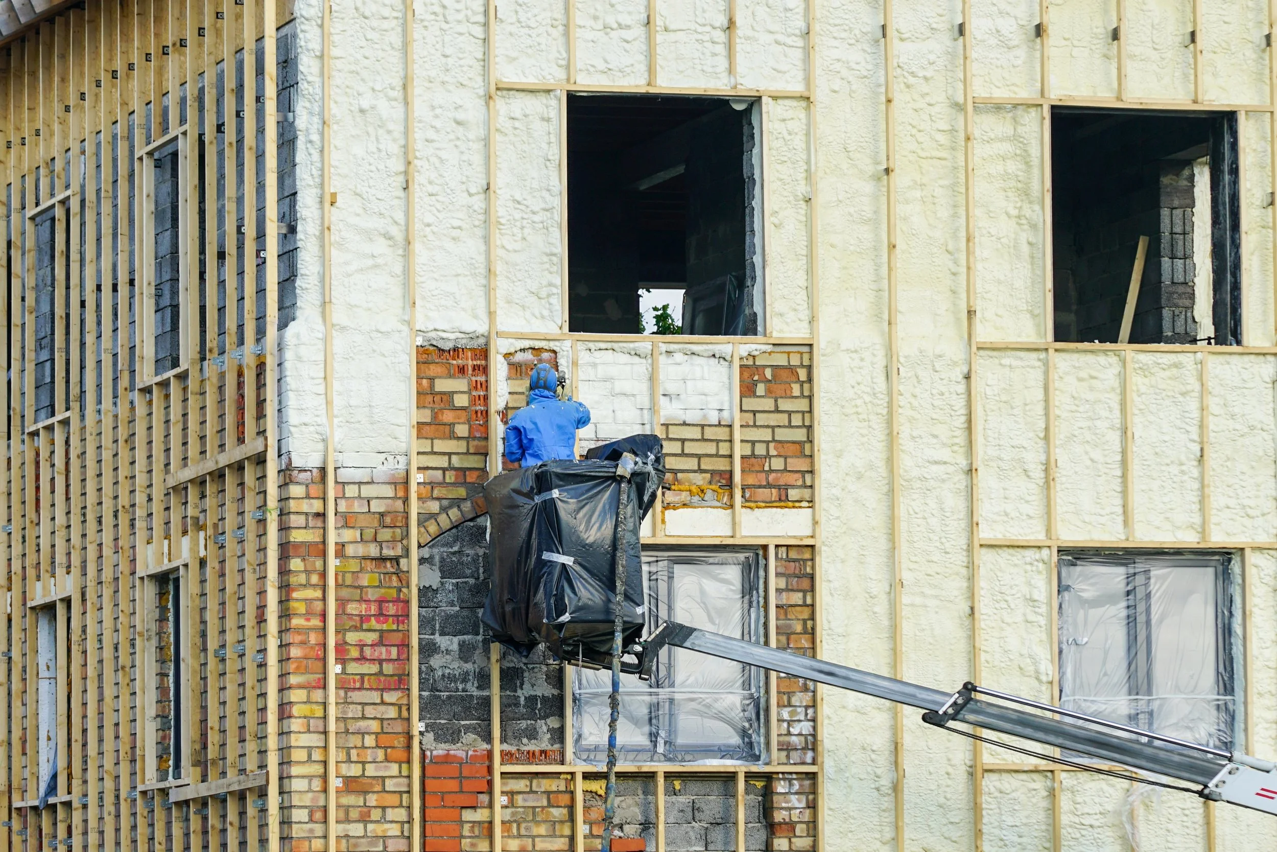 Construction worker in blue jacket and helmet operating a lift on the exterior of a building under construction, with exposed wooden framing and spray foam insulation.