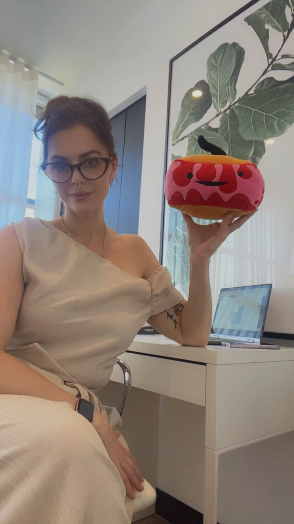 Woman with glasses sitting at a desk, holding a plush toy shaped like a smiling pumpkin with a black stem, in front of a window and a plant-themed wall art.