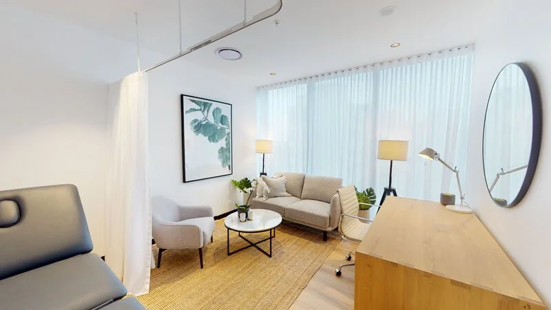 Bright, modern living room with white walls, large window with sheer curtains, beige sofa, white armchair, round coffee table, wooden desk with a circular mirror, and various potted plants.