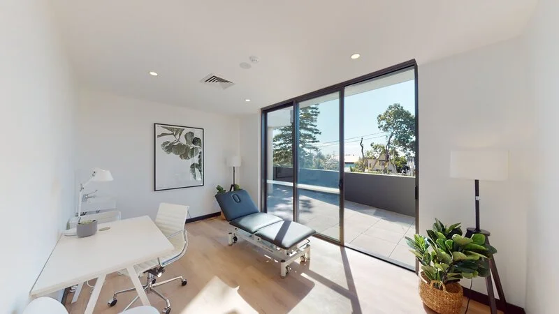 Modern medical examination room with a treatment table, desk, chair, and large window overlooking trees and a balcony.