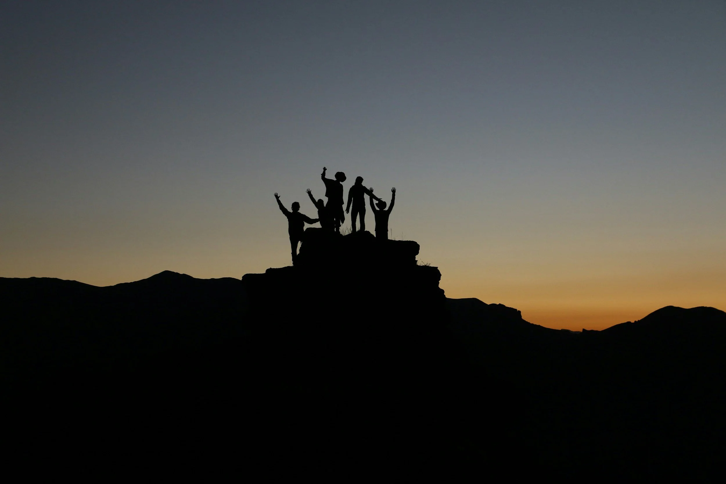 Silhouettes of five people celebrating atop a rocky hill during sunset.