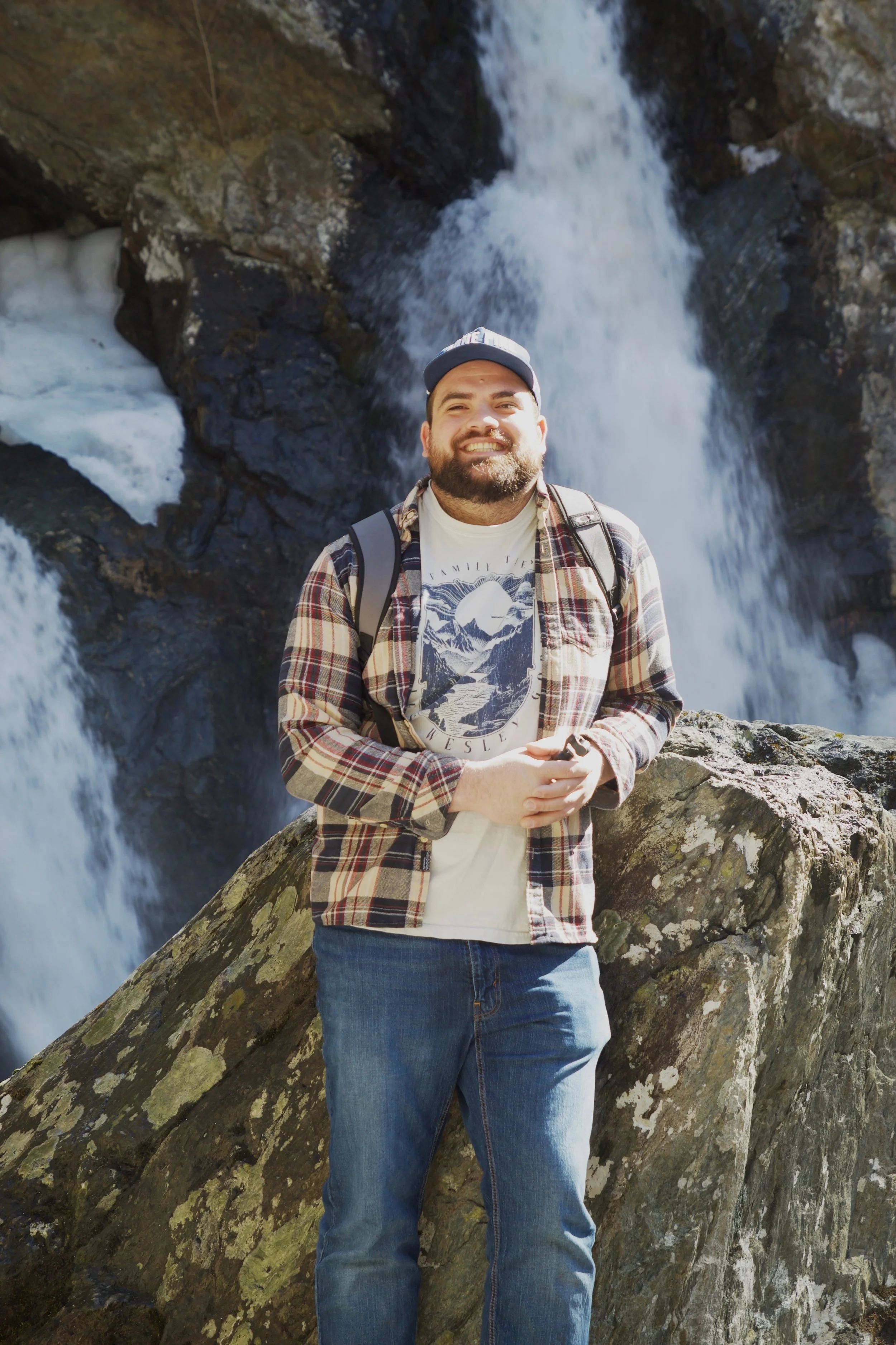 Man smiling in front of a waterfall, wearing a cap, plaid shirt, and carrying a backpack