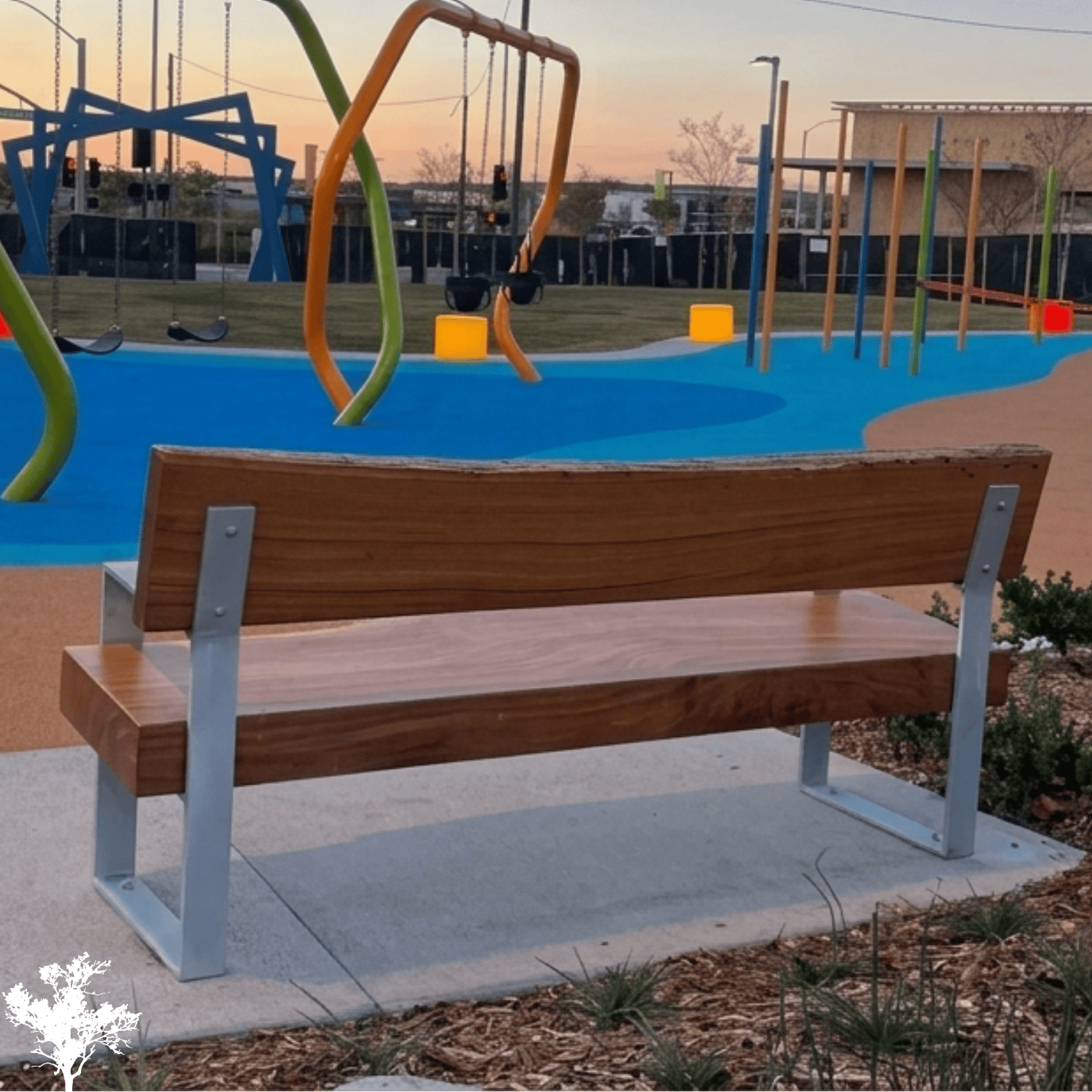 Park bench in front of a playground with swings, a slide, and climbing structures at sunset.