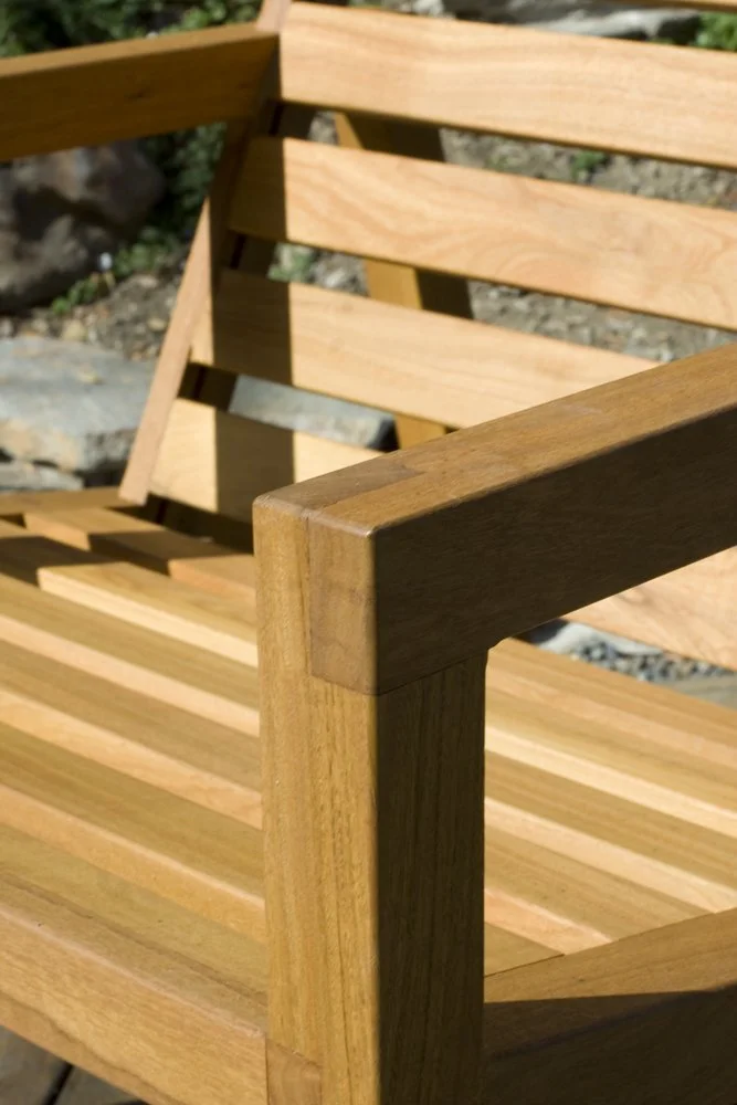 Close-up of a wooden outdoor bench with horizontal slats and armrest, with rocks and plants in the background.