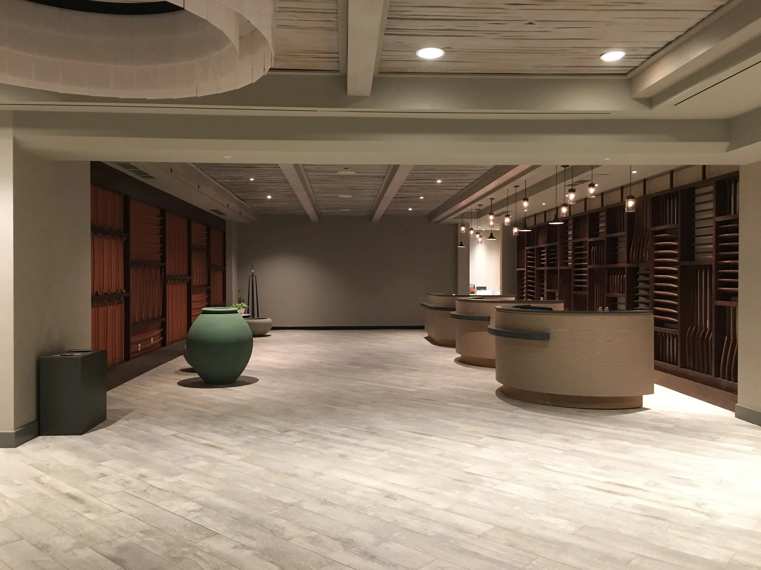 Empty hotel lobby reception area with beige curved counter, wooden wall with open shelving, green vases, and modern ceiling lighting.