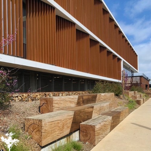 Exterior view of a modern building with a brown slatted facade, large windows, and an outdoor seating area with massive wooden blocks on a cement pathway, under a partly cloudy sky.