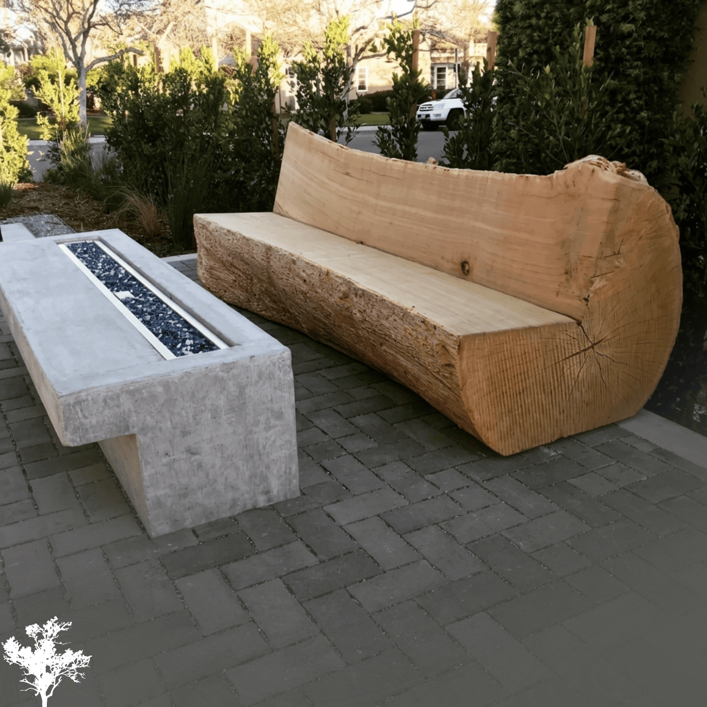 A large wooden bench with a natural, live edge design and a rectangular concrete fire pit with a black gravel center, set on a paved outdoor area with greenery and trees in the background.