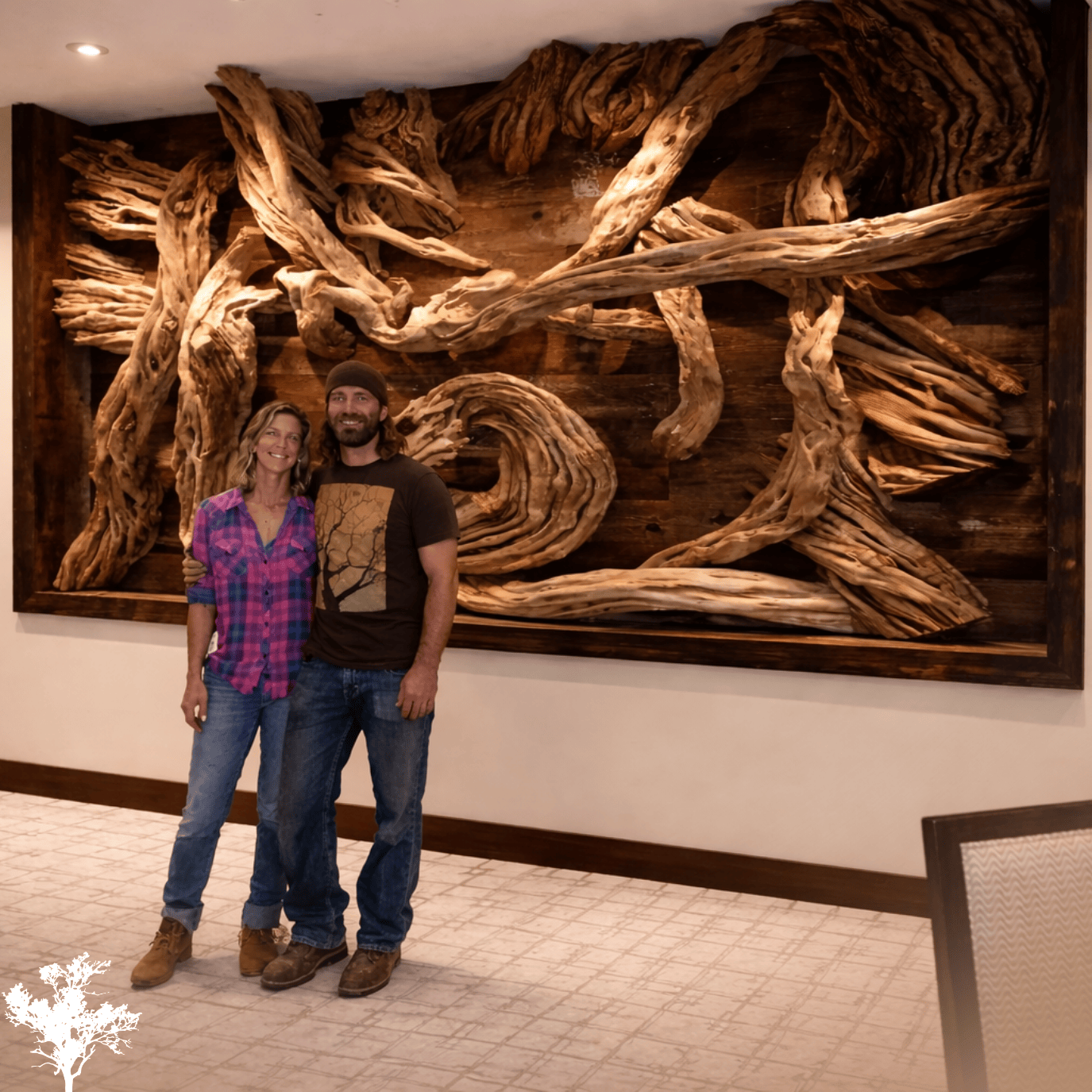 A woman and a man standing together in front of a large, intricate wooden art piece mounted on a wall indoors. The woman is wearing a pink and purple plaid shirt and jeans, while the man is dressed in a brown T-shirt with a rectangular graphic and jeans. They are both smiling, with their arms around each other.