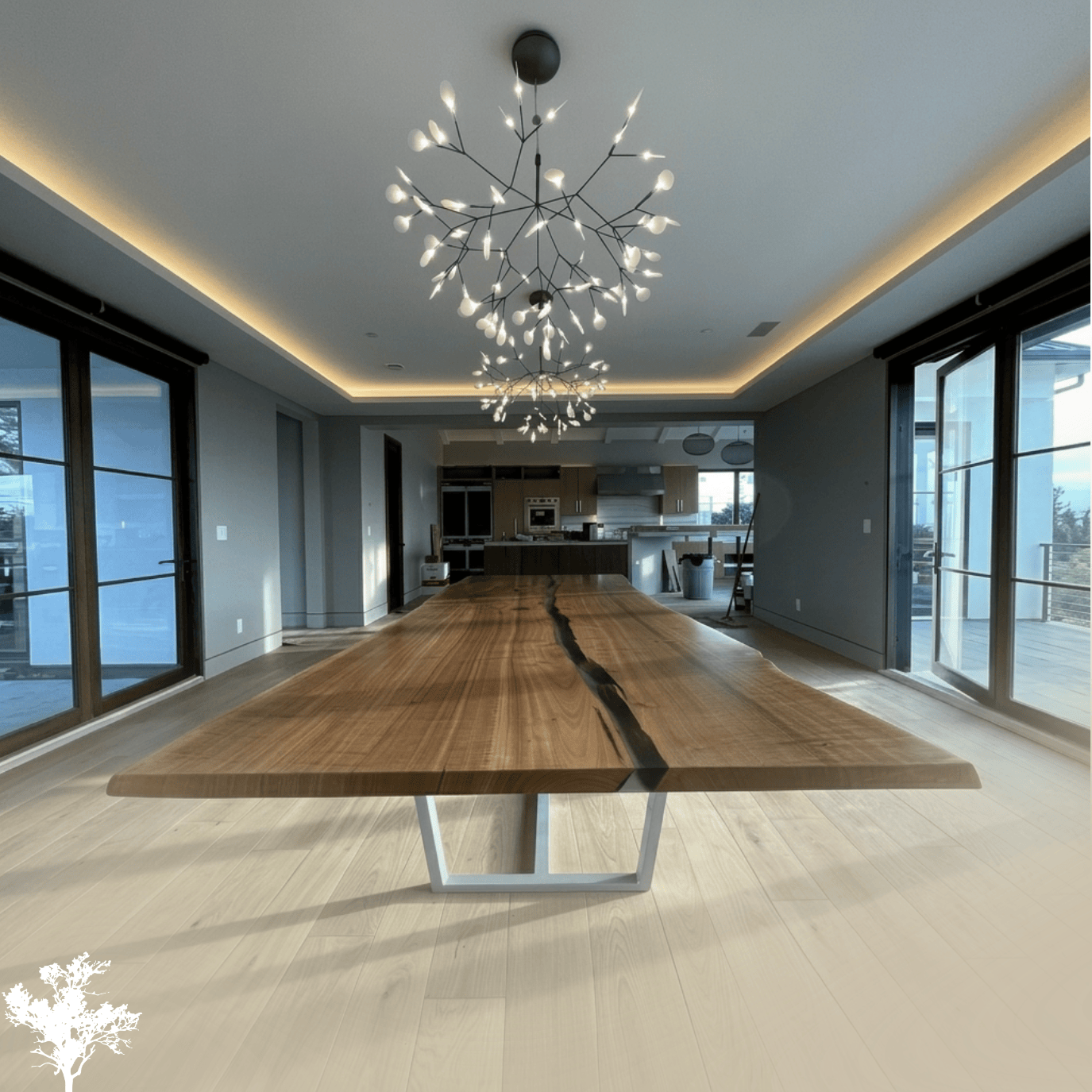 Modern dining room with large wooden table, black branch-like chandeliers, and sliding glass doors leading to balcony, open kitchen in background.