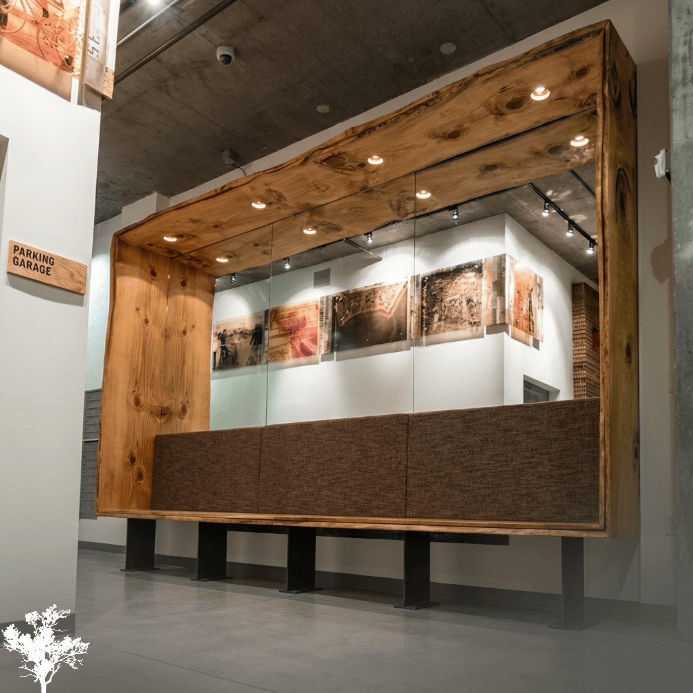 A modern interior with a large wooden and glass display booth with brown fabric seating underneath, in an art gallery or gallery-like space. White walls, concrete ceilings, track lighting, and artwork are visible in the background.