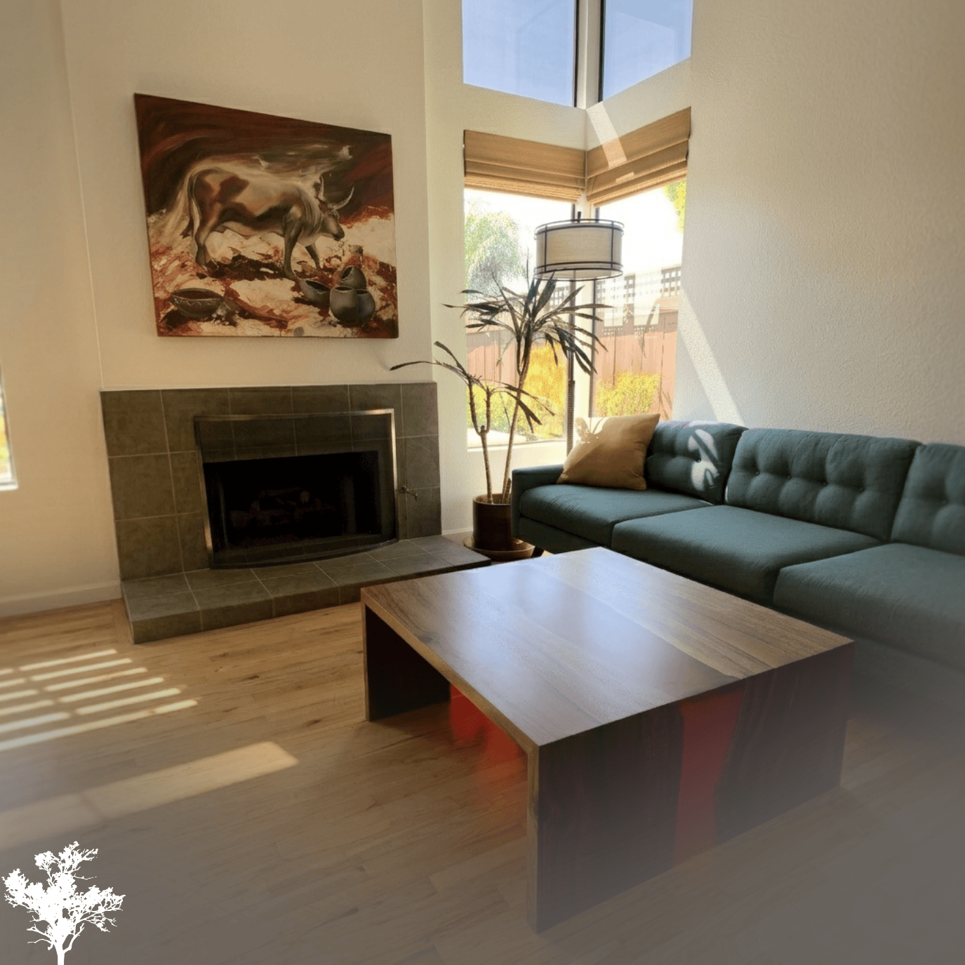 Living room with a fireplace, a green sofa with a beige pillow, a wooden coffee table, a potted plant, a floor lamp, large windows with beige blinds, and a painting of a bull on the wall.