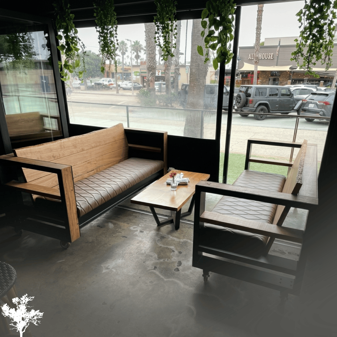A cozy seating area in a cafe featuring a wooden bench and a matching armchair with leather cushions, a small wooden coffee table with flowers, and green hanging plants above. Large windows reveal a street with parked cars and palm trees outside.