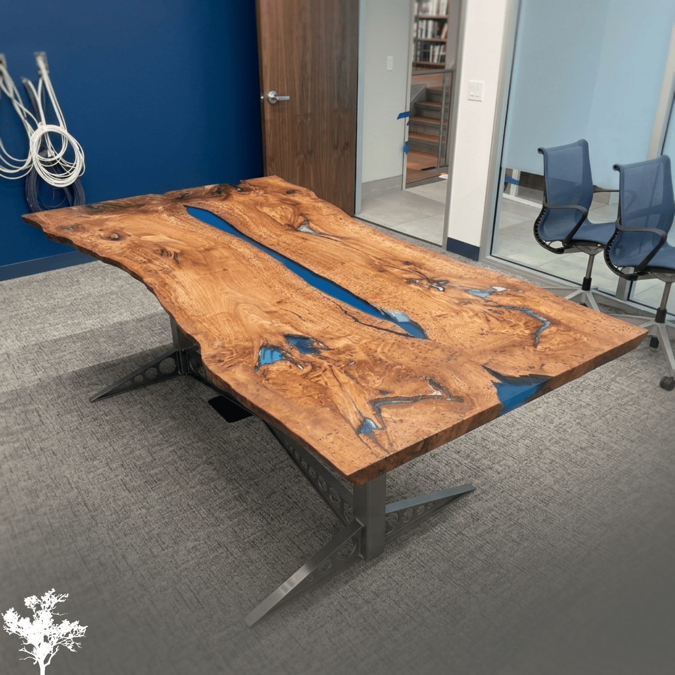 A large wooden conference table with a natural, irregular shape and a central blue resin inlay in a modern office conference room.