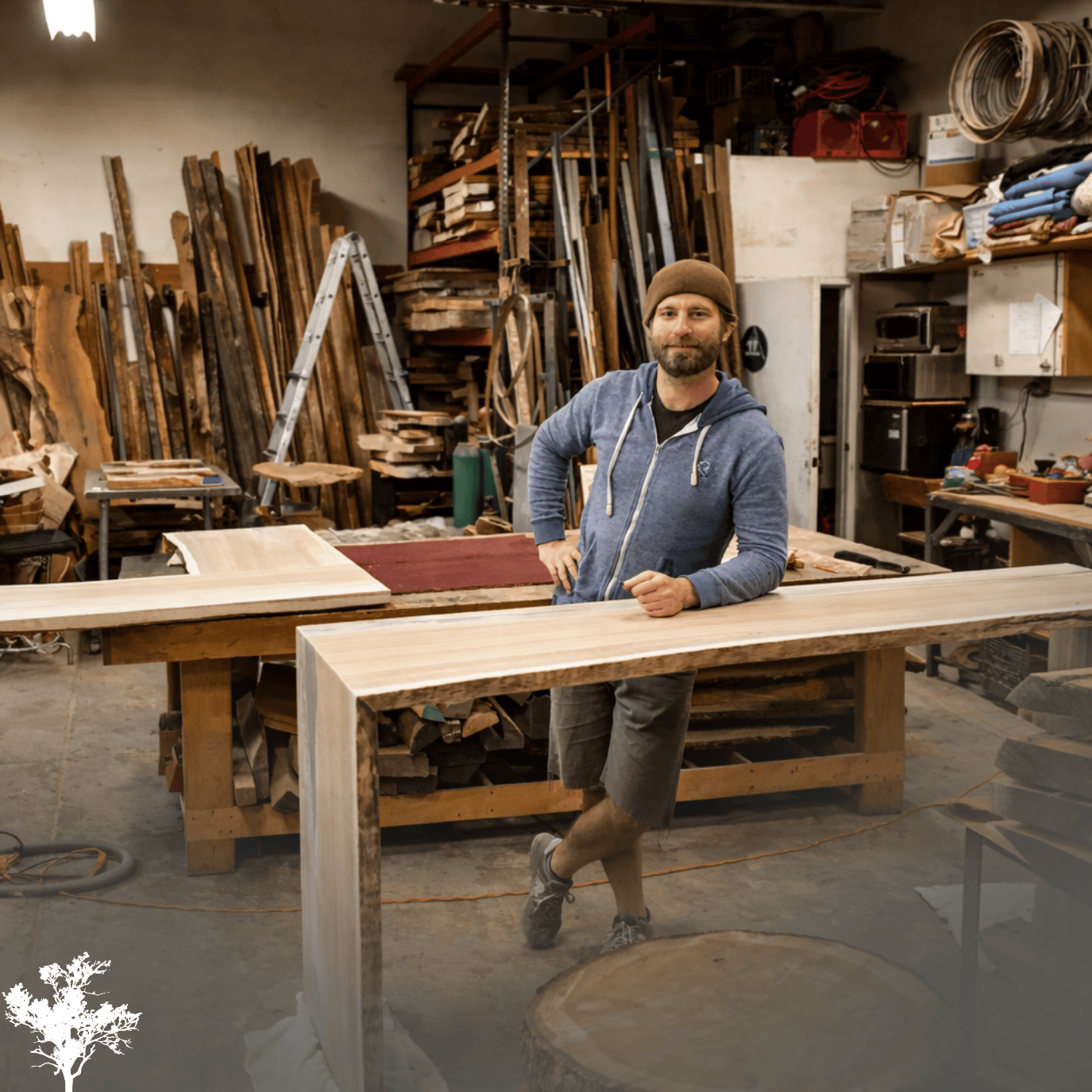 A man in a workshop surrounded by wooden planks and woodworking tools, standing beside a long piece of wood on a work table.