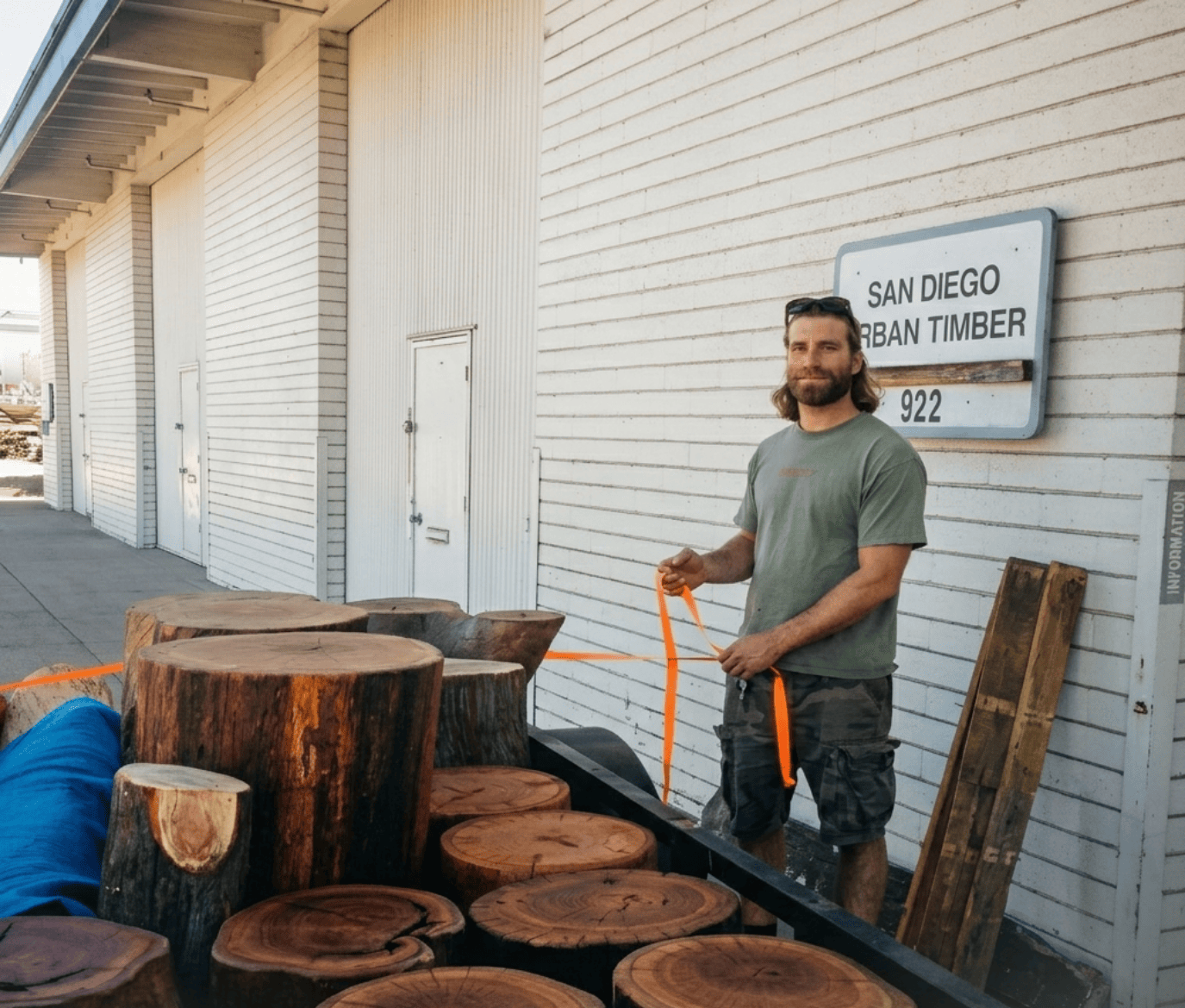 A man with shoulder-length hair and a beard, wearing a green t-shirt and camouflage shorts, stands outside next to a white building with a sign that reads 'San Diego Urban Timber'. He is holding an orange strap tied around large cut logs. The logs are stacked in the bed of a trailer or truck.