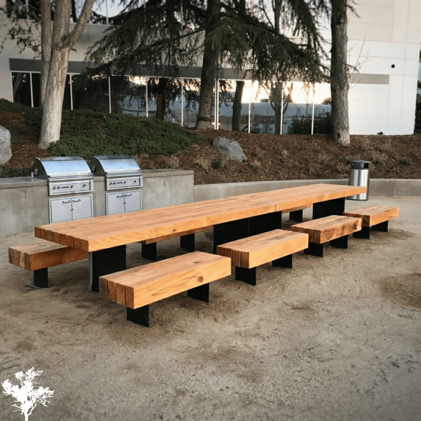Wooden picnic table with attached benches and stainless steel trash and grill stations in an outdoor park area with trees and a building in the background.