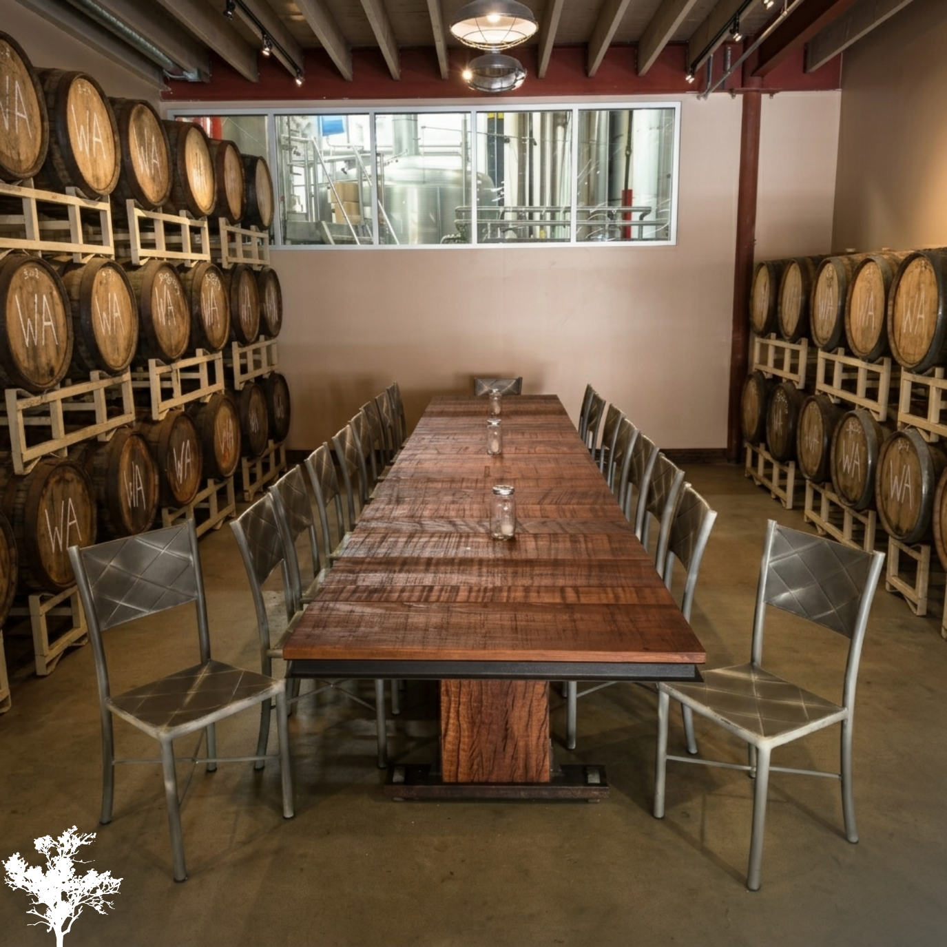 A long wooden table surrounded by metal chairs in a wine cellar, with large wine barrels stacked against the walls and a window looking into the winery equipment.
